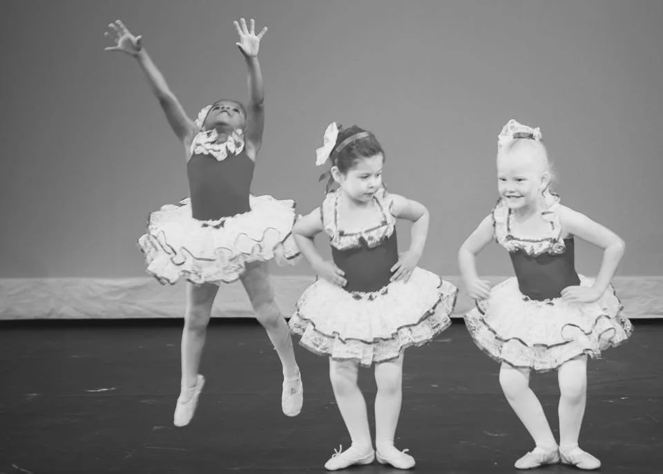 Three young girls dressed in ballet costumes with tutus and bows, performing a dance on stage. The girl on the left is jumping with arms raised, and the other two are standing with hands on hips, smiling.