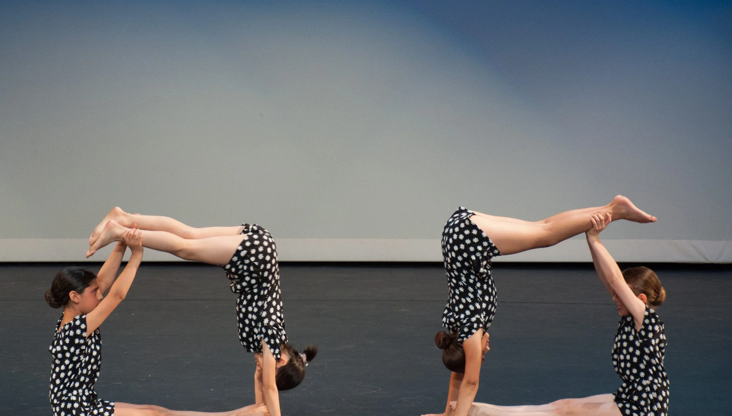 Two pairs of young girls dressed in matching black and white polka dot outfits perform yoga or acro yoga on stage, with one girl in each pair balancing on the other's hands and feet in a headstand pose.