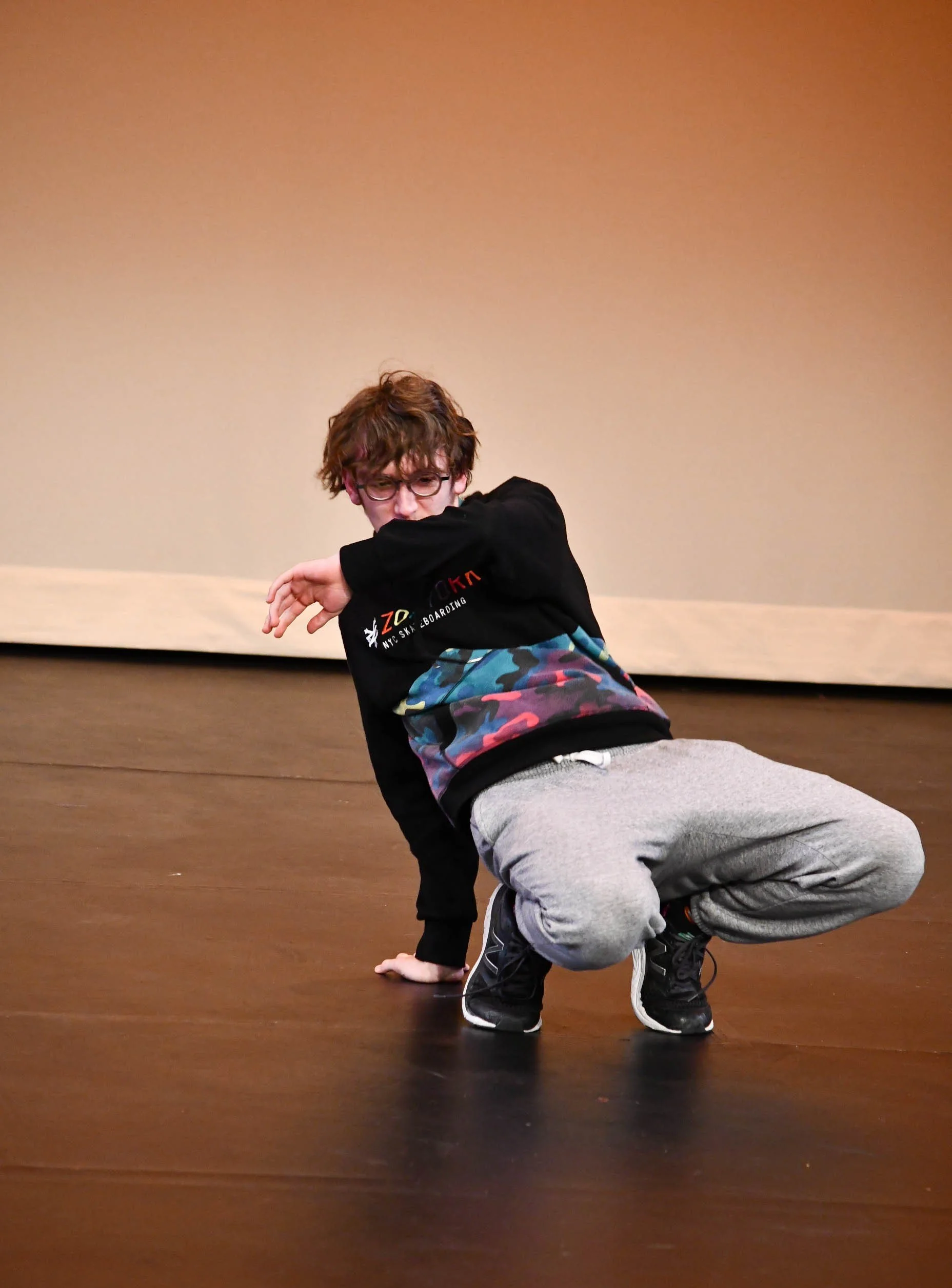 A young person with curly brown hair and glasses performing a dance move on a dark wooden floor with a plain beige wall background.