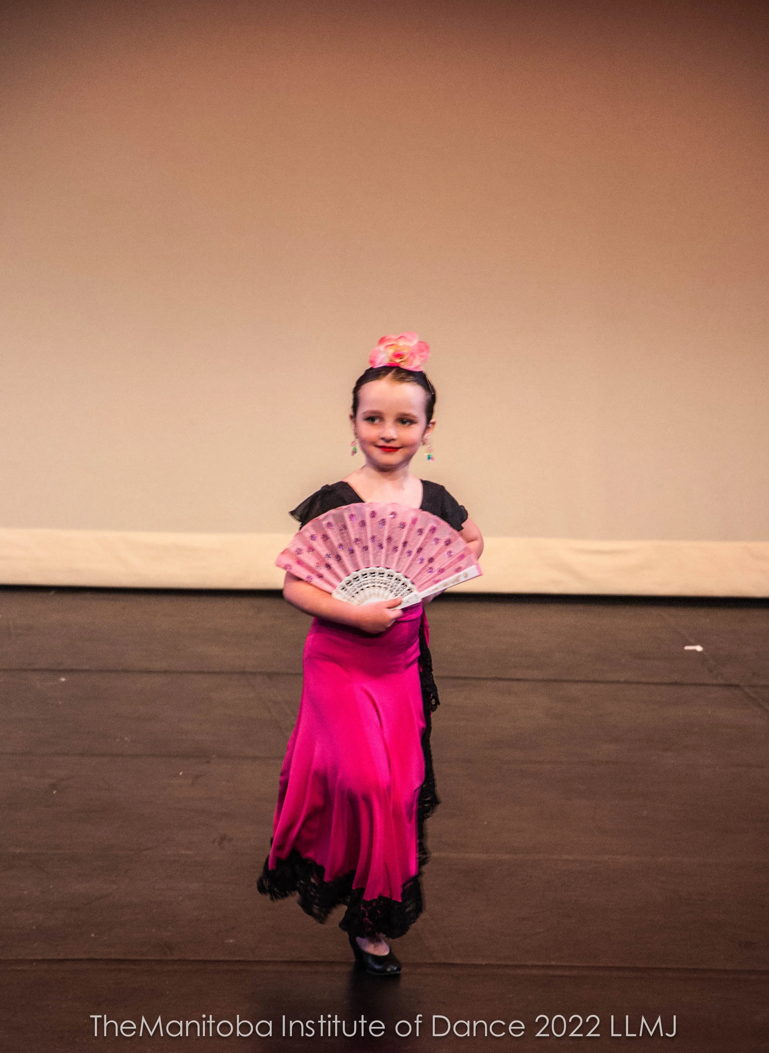 Young girl performing a flamenco dance on stage, wearing a pink and black dress, holding a pink fan, with a pink flower accessory in her hair, at The Manitoba Institute of Dance 2022.
