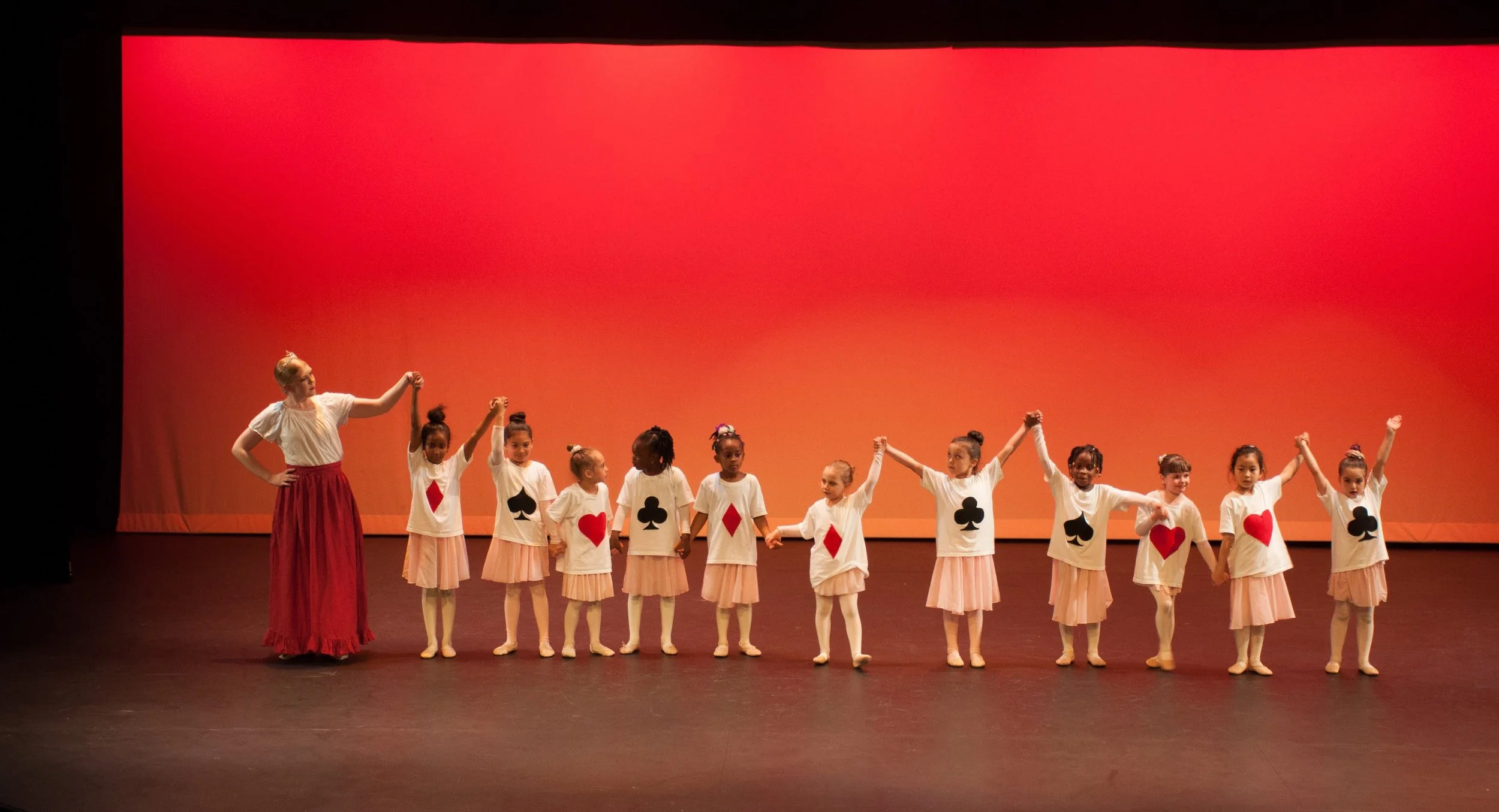 A group of young girls and an adult woman on stage, holding hands, dressed in costumes inspired by playing cards, with card suit symbols on their shirts, during a dance performance against an orange backdrop.