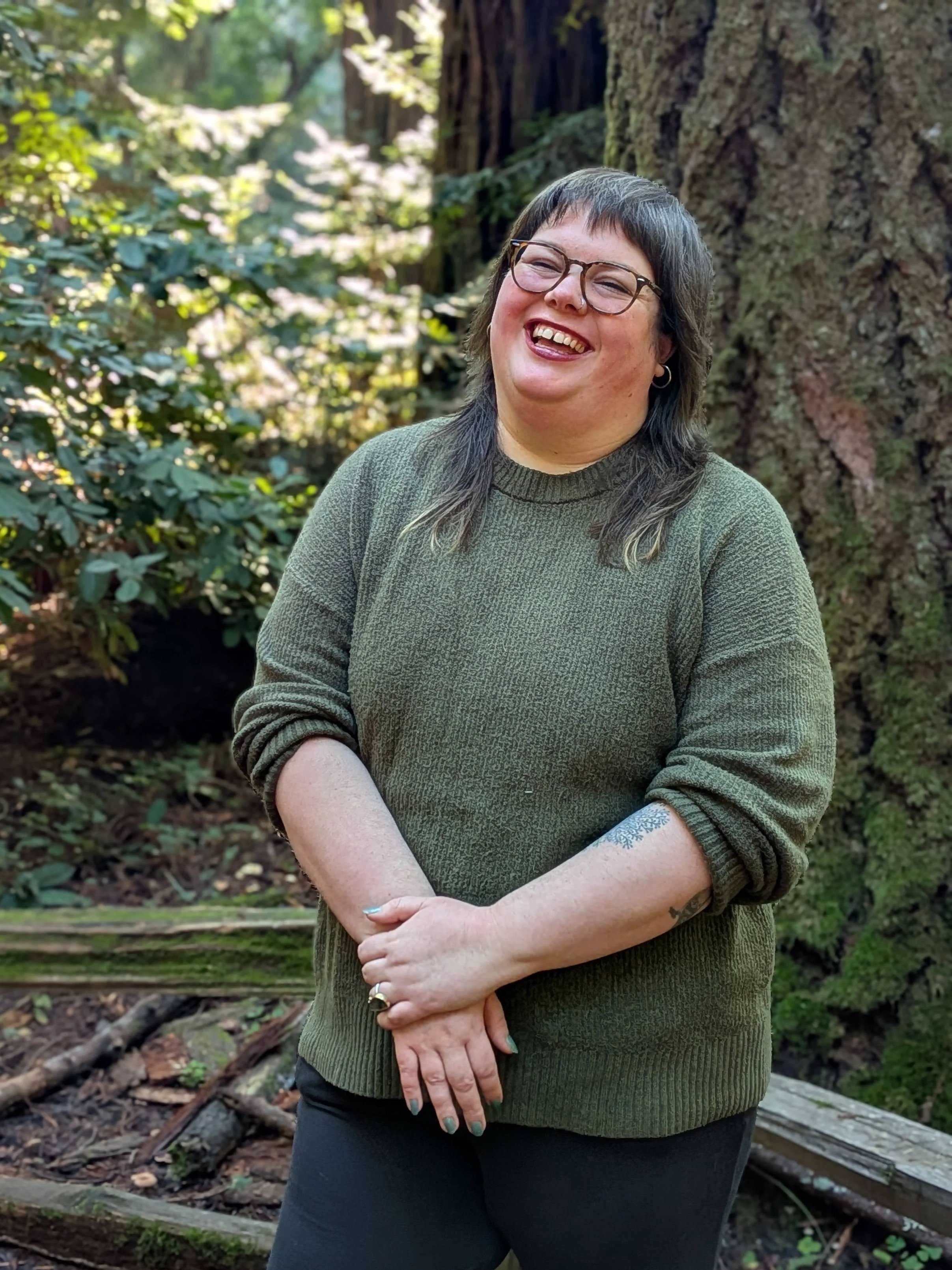 A woman with dark hair and glasses wearing a green sweater standing outdoors in a forest near a large tree, smiling.