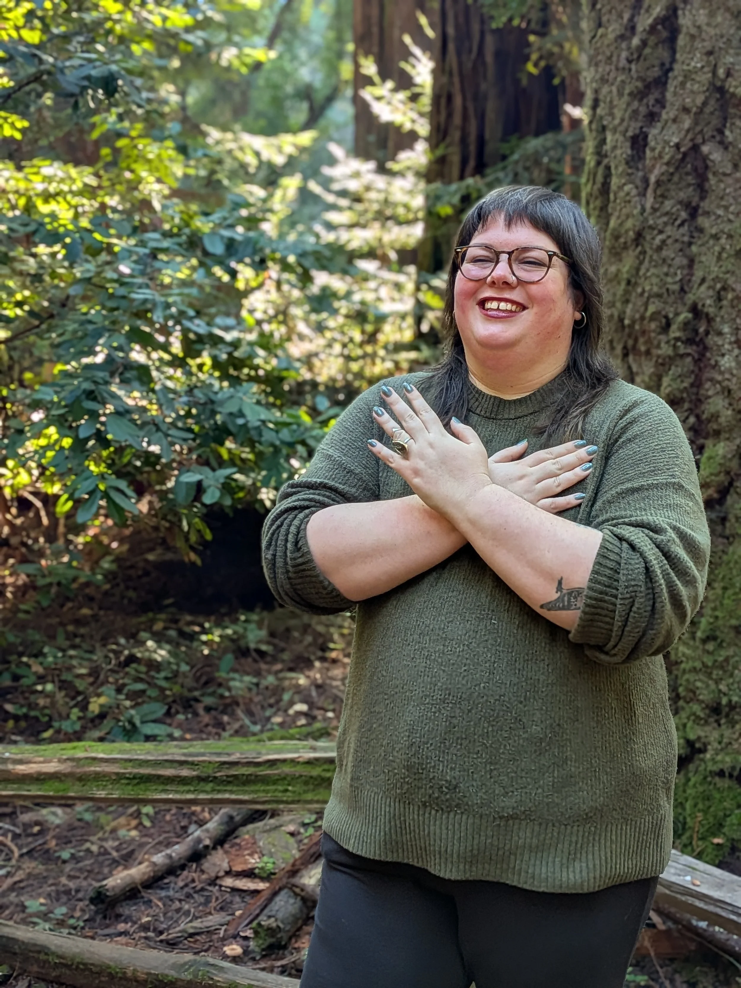 A smiling woman with glasses and long dark hair standing in a forest, crossing her arms over her chest with her hands on her shoulders, wearing a green sweater and black pants.