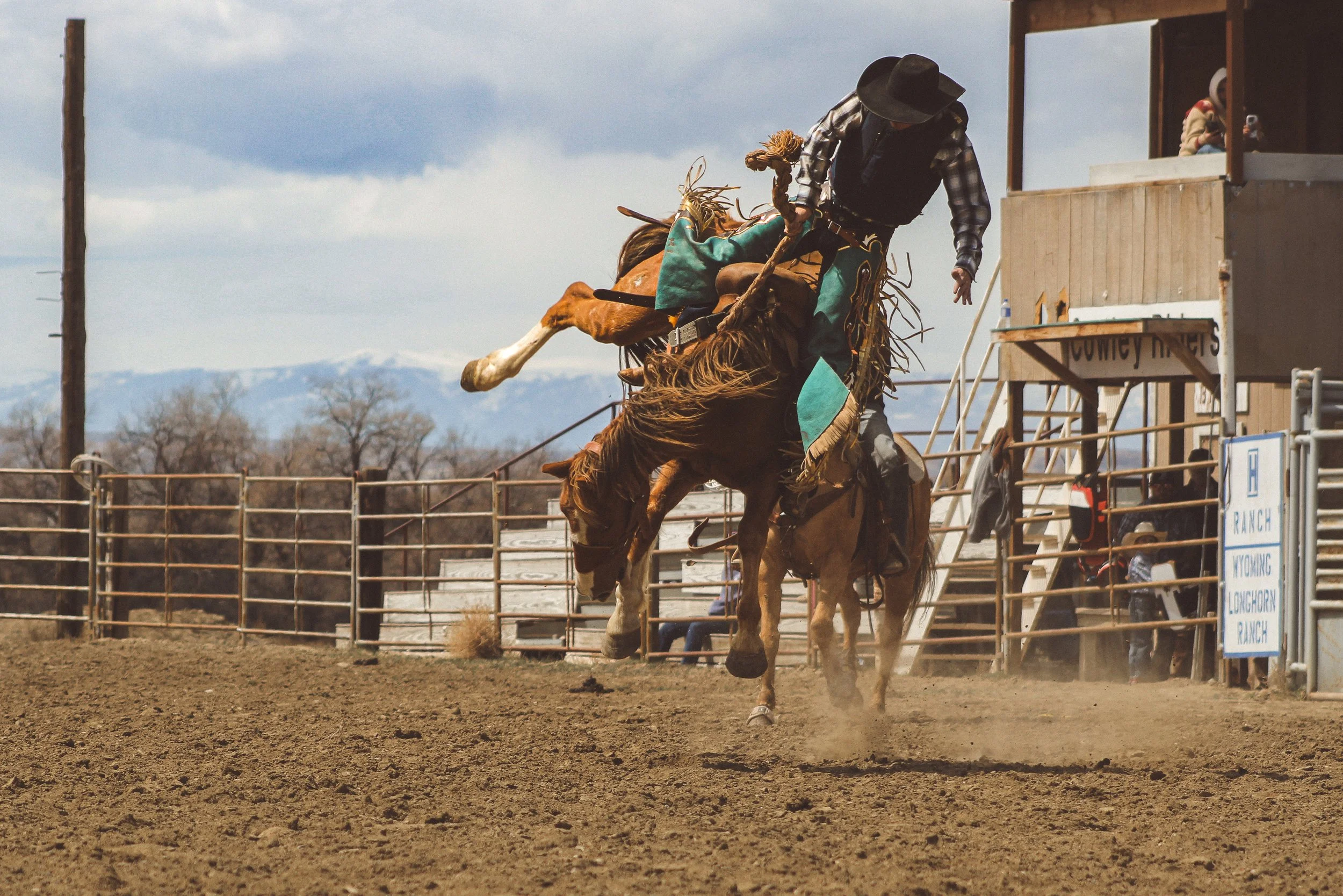 Cowboy riding a bucking horse at a rodeo arena with a dirt ground, wooden fence, and spectators in the background.