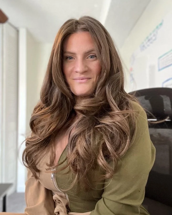 A woman with long, wavy brown hair sitting in an office chair, smiling softly at the camera, with a white wall and some colorful posters or charts in the background.