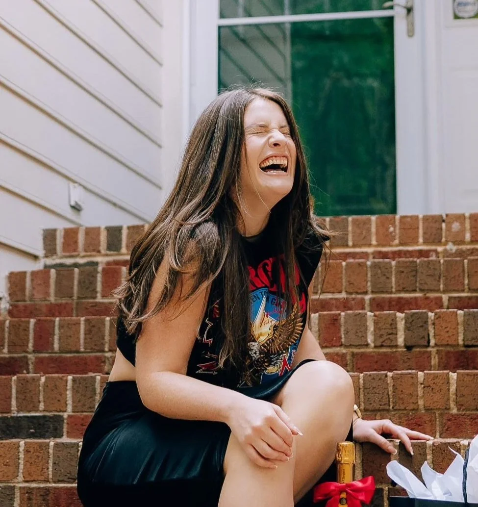 A woman with long brown hair laughing on brick stairs outside a house, wearing a black sleeveless shirt with a graphic print.