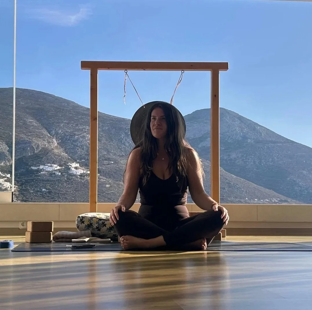 Woman practicing yoga indoors on a mat, sitting cross-legged, with a mountain landscape visible through a large window behind her.