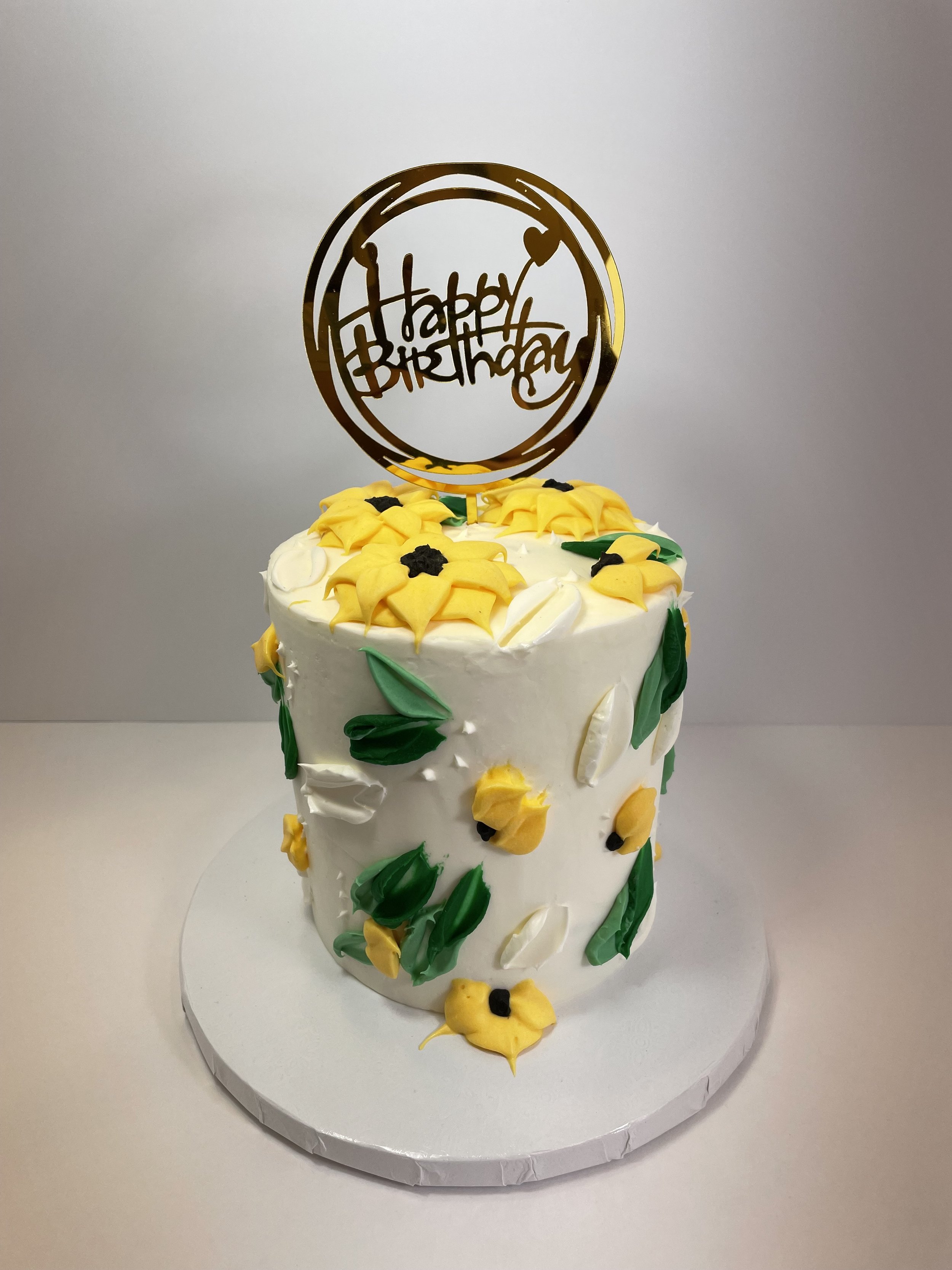 A white birthday cake decorated with yellow flowers, green and white leaves, topped with a gold 'Happy Birthday' topper.