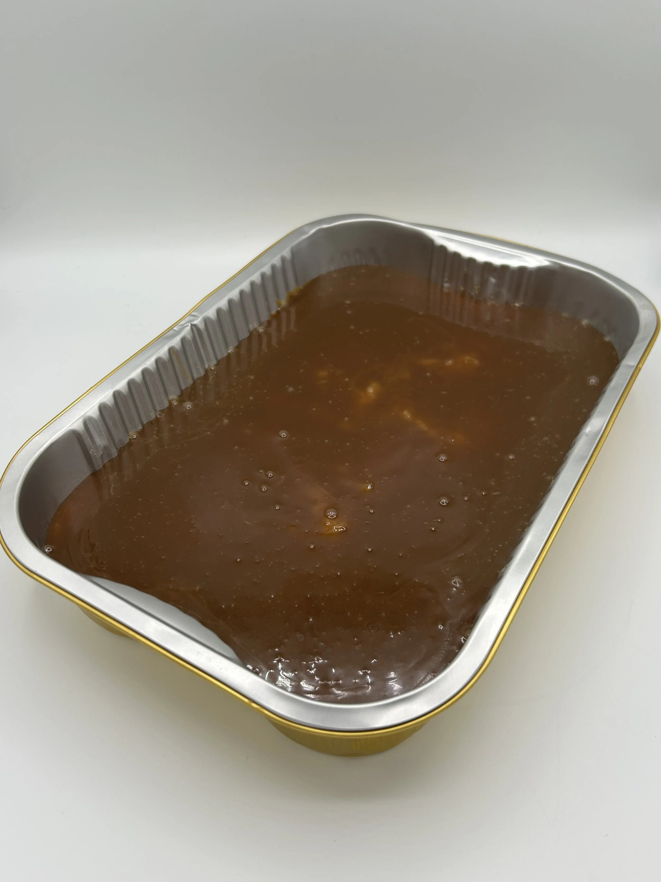 A rectangular aluminum tray filled with brown gravy or sauce, placed on a white surface. Biko