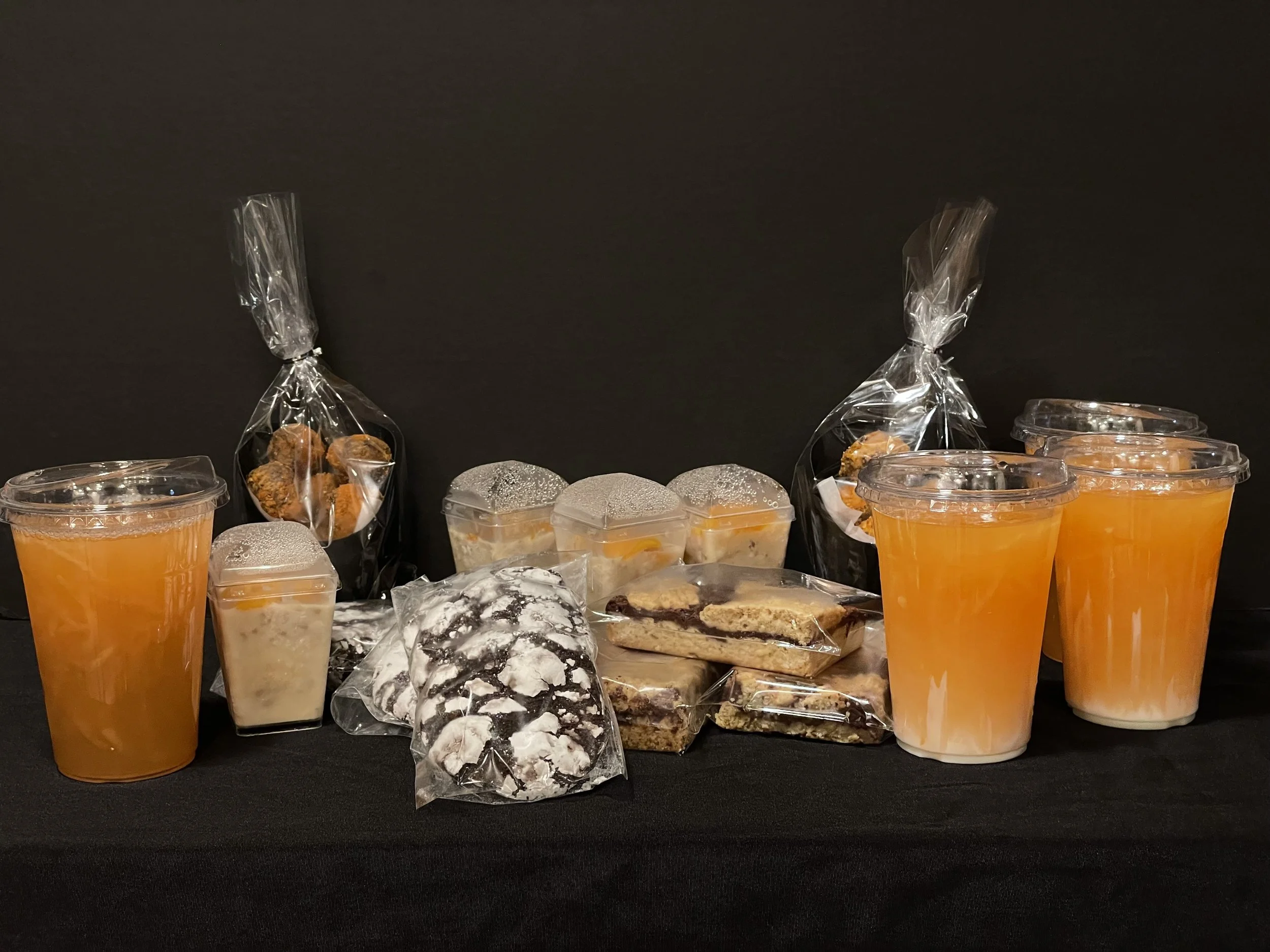 Assorted baked goods and beverages on a black surface, including cookies, cakes in cups, sealed sandwiches, and cups of orange drink.