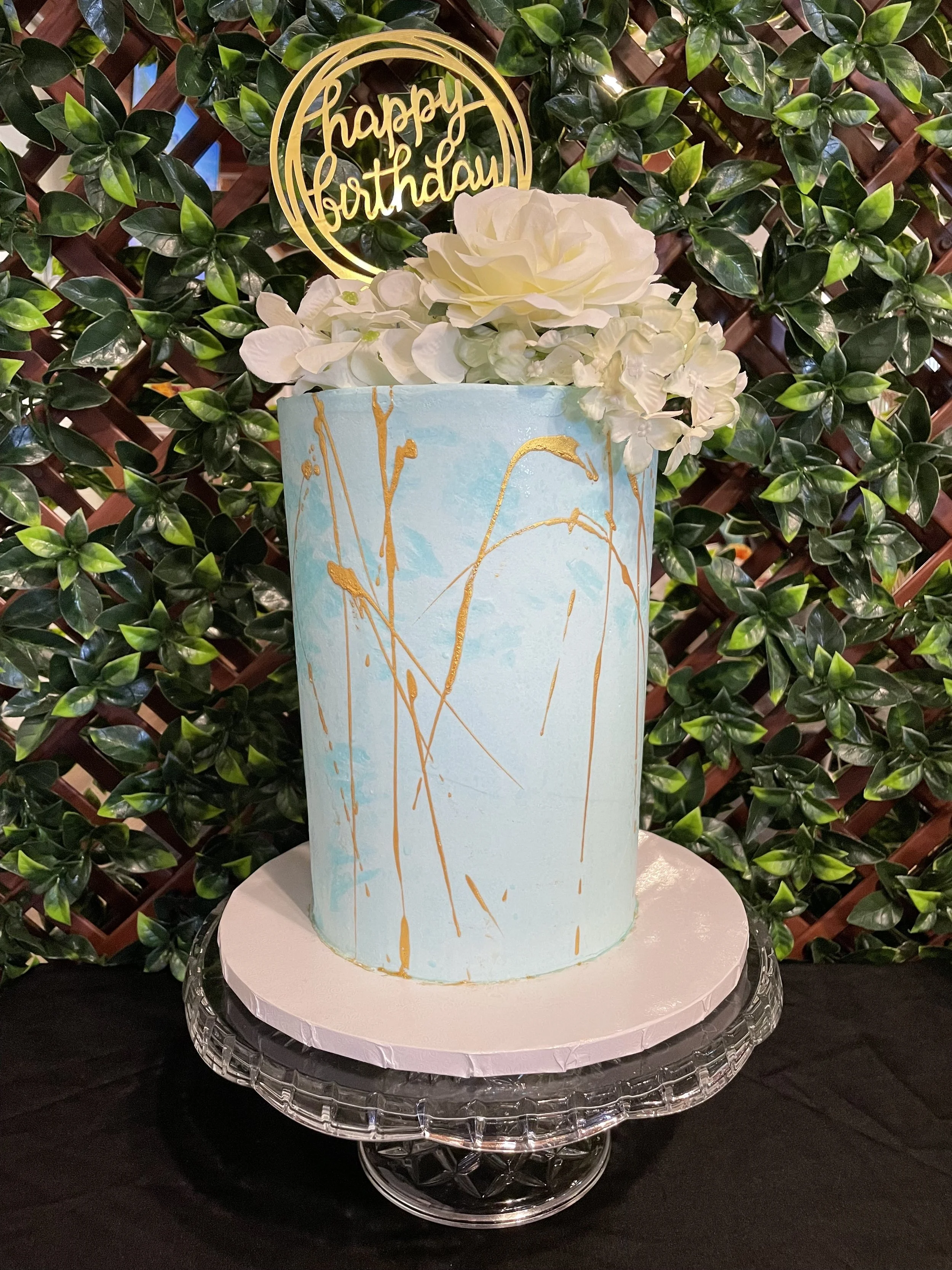 A tall birthday cake with watercolor blue and gold splatter design, decorated with white flowers on top, and a gold "happy birthday" topper, displayed on a glass cake stand with a black cloth surface and green leafy background.