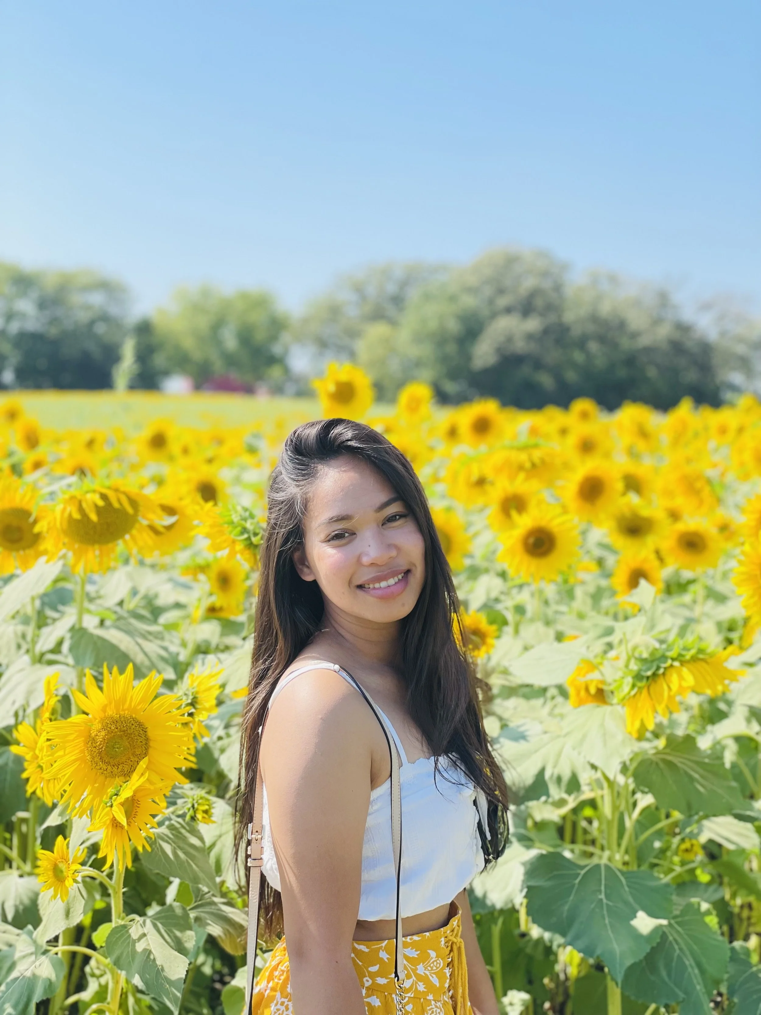 A woman with long dark hair smiling in a sunflower field on a bright sunny day with a clear blue sky and trees in the background.