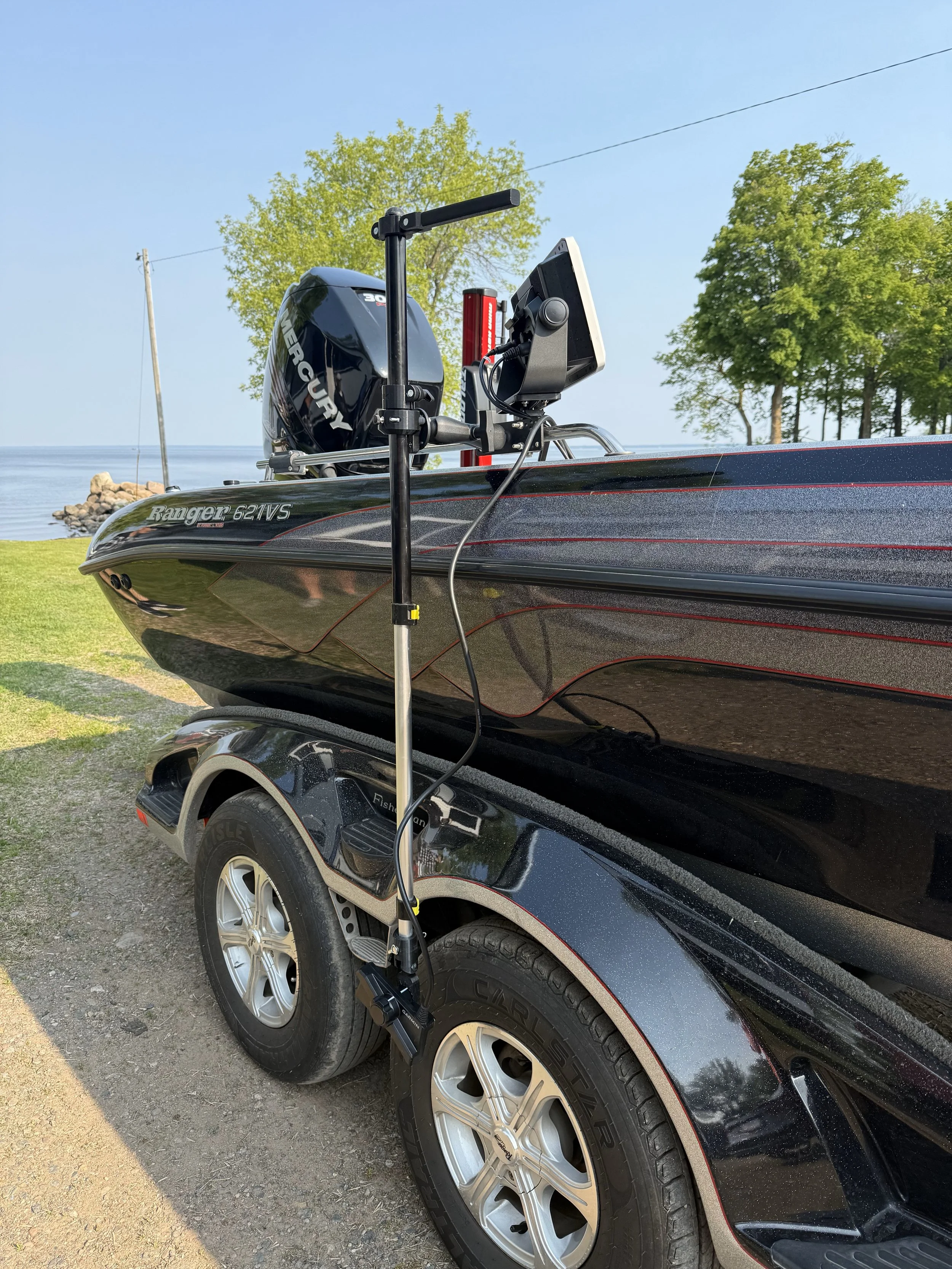 A black Ranger 621VS fishing boat on a trailer with a Mercury outboard motor, parked on a gravel surface near a lake with trees and a small dock in the background.