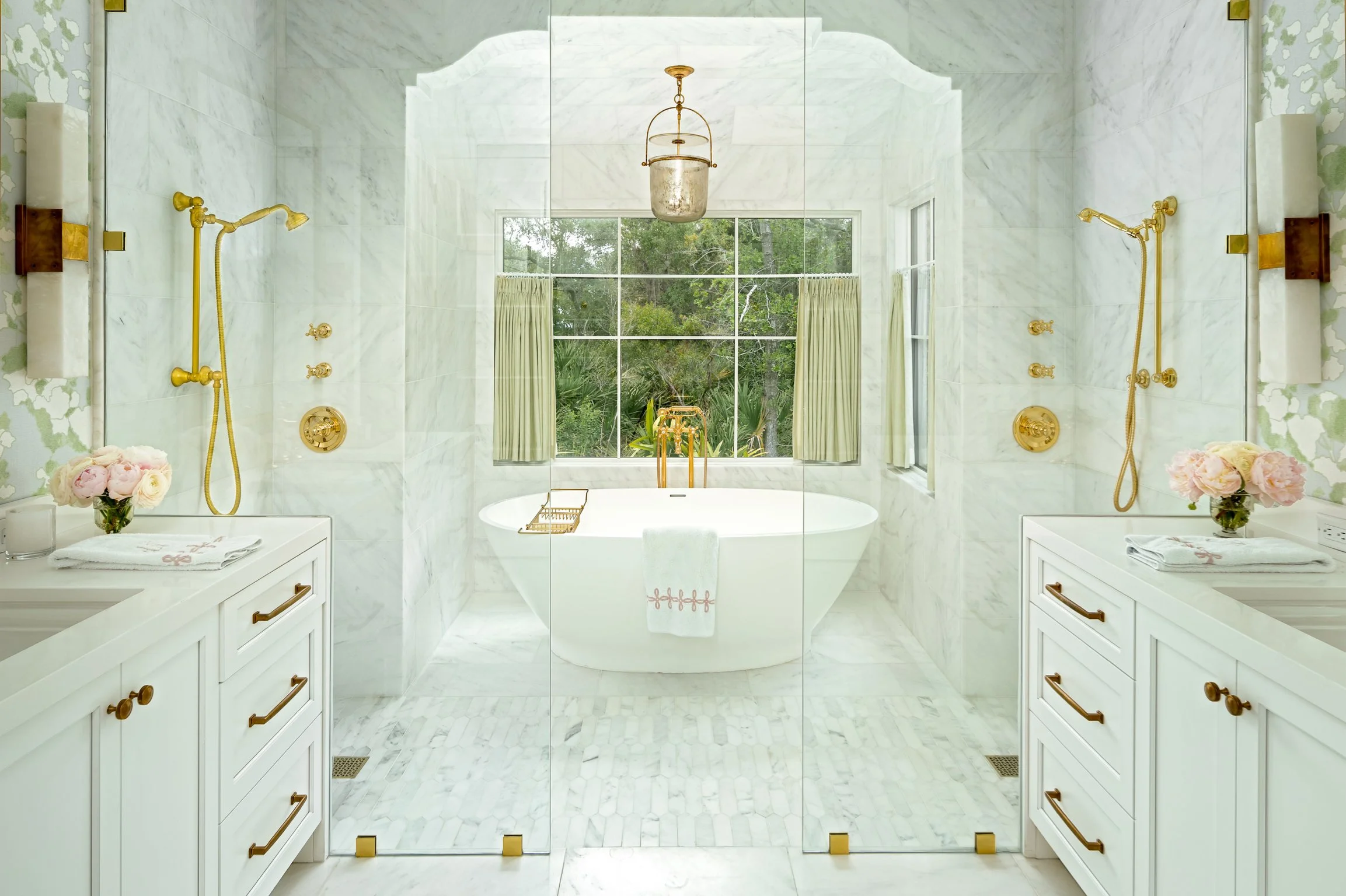 A luxurious bathroom with a large window revealing trees outside. The bathroom features white marble walls and floor, a freestanding bathtub, two white vanities with gold fixtures, and gold showers. A central chandelier hangs from the ceiling.