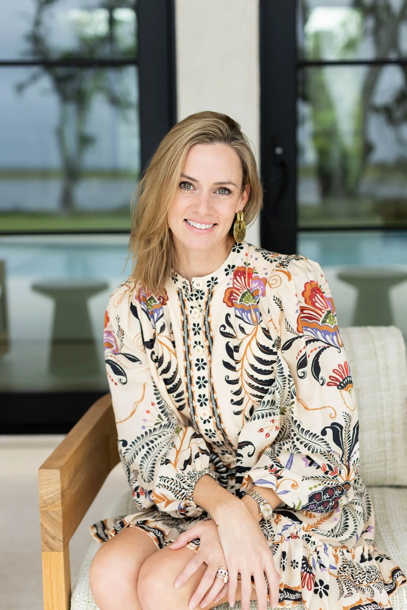 A woman with blonde hair and earrings sitting on a wood chair in front of large glass windows, smiling at the camera, wearing a multicolored patterned dress.