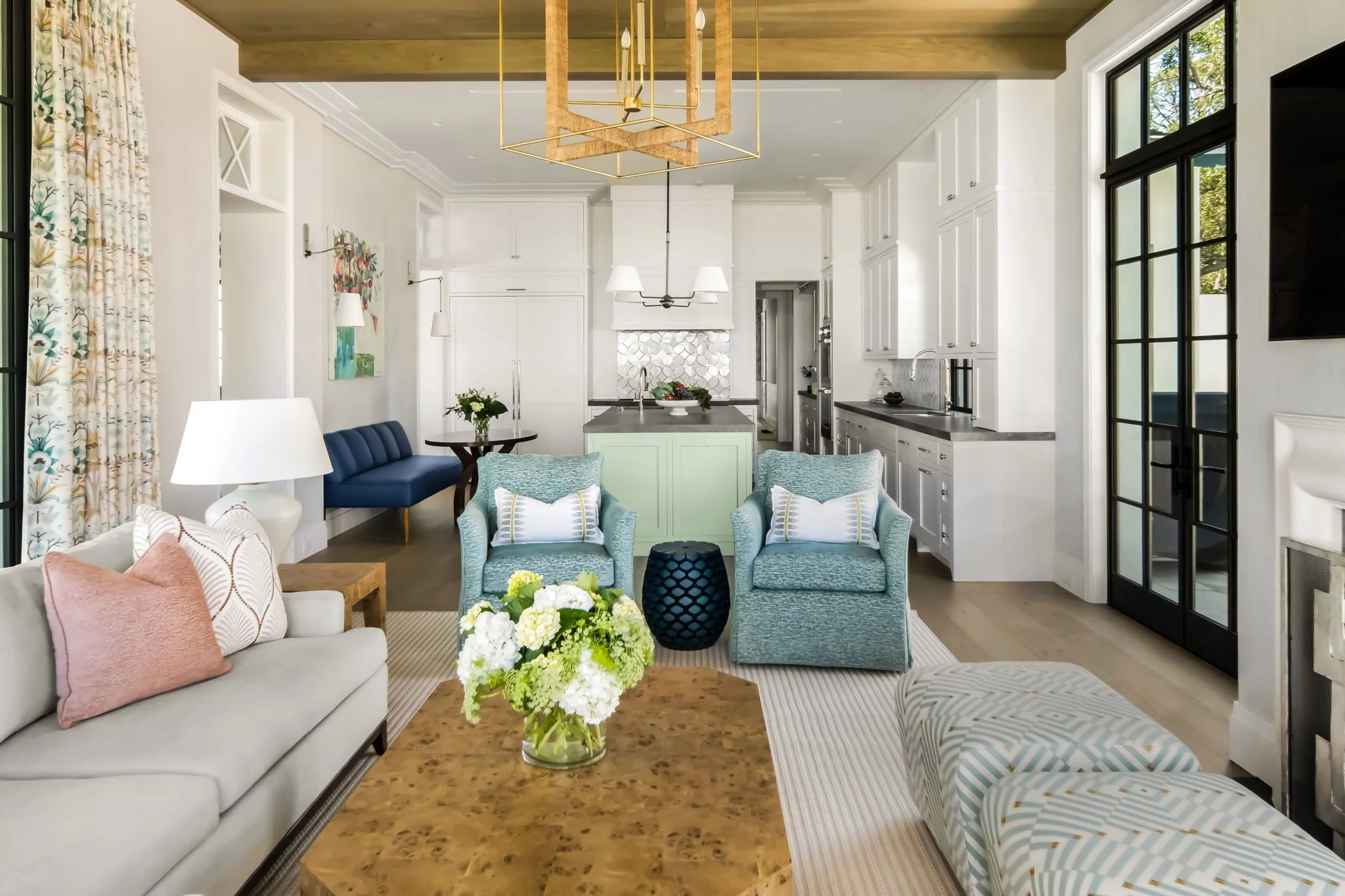 A bright and airy living room with white walls, a light wood ceiling, and large windows with black frames. The room features a white sofa with pink and patterned pillows, two light blue armchairs with striped pillows, a wooden coffee table with a floral arrangement, and a dark blue pouf. There is a navy built-in bench with a dark blue cushion, lamps, and wall art. The kitchen area in the background has white cabinetry, a green kitchen island, and modern lighting fixtures.