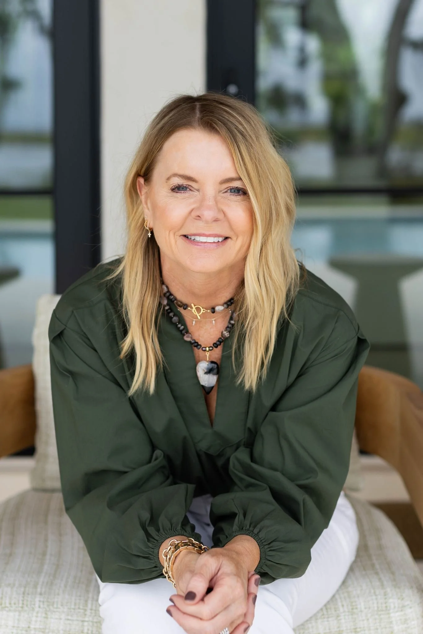 A middle-aged woman with blonde hair sits on a bed, smiling, wearing a green long-sleeve blouse and jewelry, with a large window in the background.