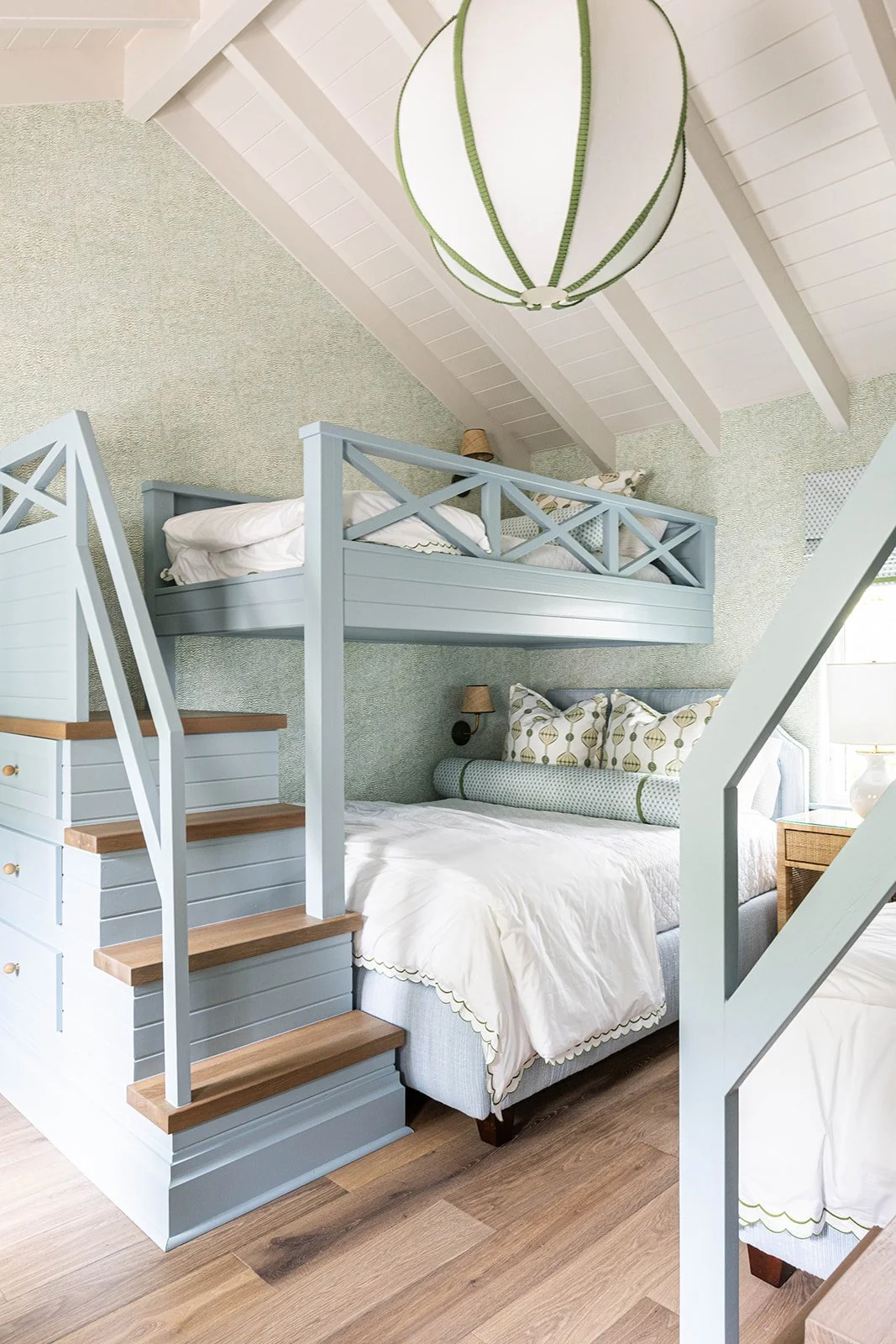 A cozy bedroom with a bunk bed, featuring a lower full-size bed with decorative pillows and a white comforter, and an upper twin-size bed accessible by wooden stairs. The room has a light, beachy decor with white walls, a textured green wall, and a large, round light fixture hanging from a white ceiling with exposed beams.