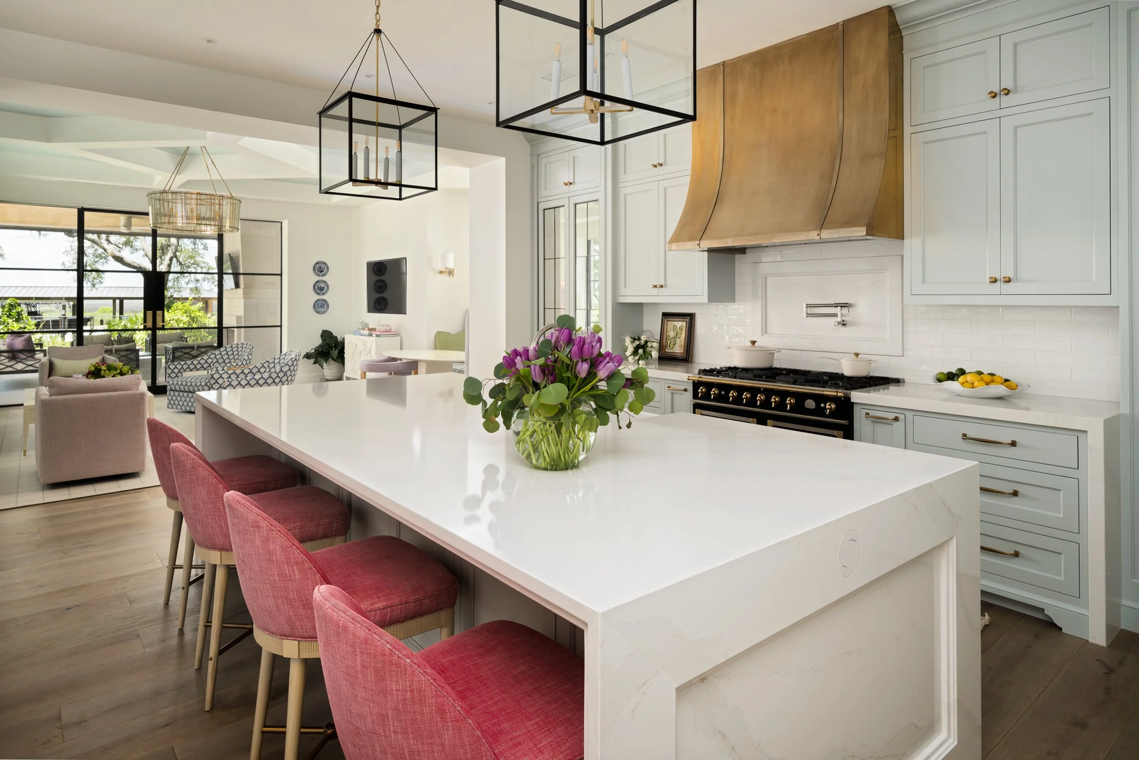 Modern kitchen and living room with white cabinetry, pink chairs around a large kitchen island, stainless steel appliances, and a vase of purple tulips on the island.