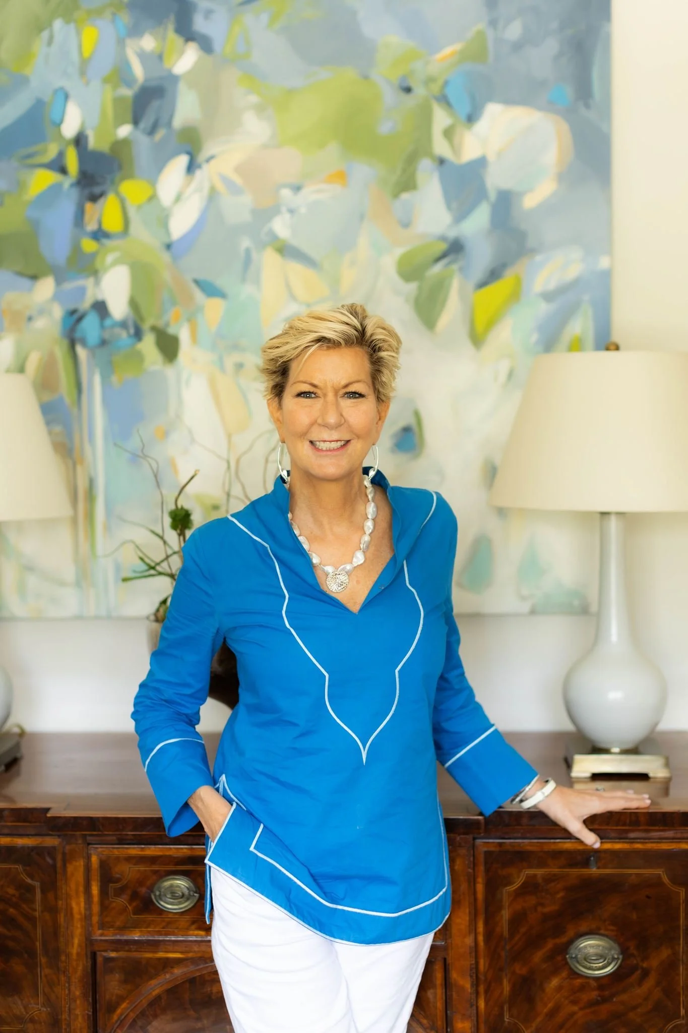 A smiling older woman with short blonde hair standing indoors in front of an abstract painting. She is wearing a bright blue top with white piping details, white pants, a shell necklace, and earrings. She has her left hand resting on a wooden cabinet and is looking at the camera.