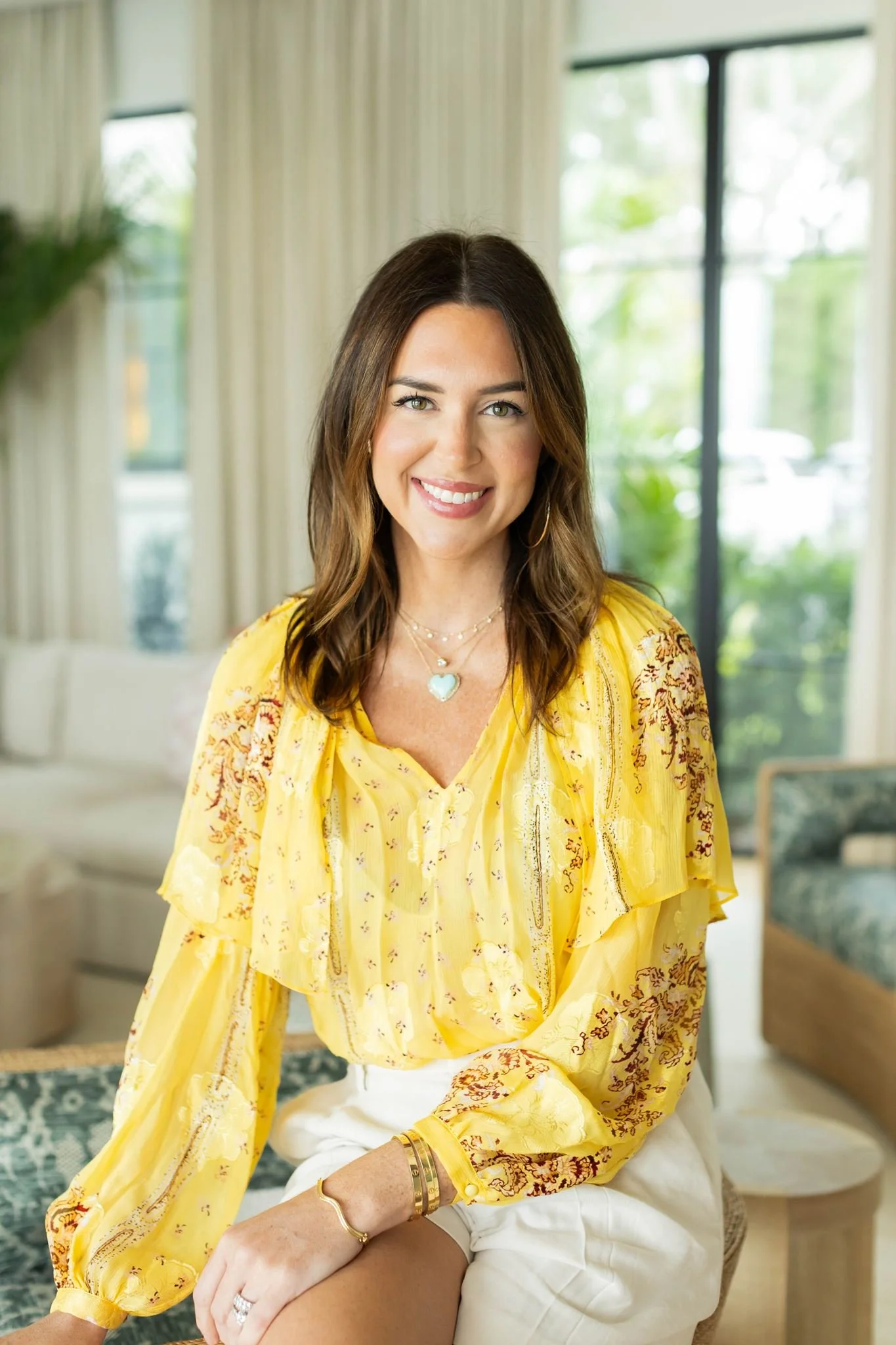 Woman with long brown hair and light eyes smiling, wearing a yellow embroidered blouse, jewelry including a blue heart necklace, sitting in a modern bright room with large windows and greenery outside.