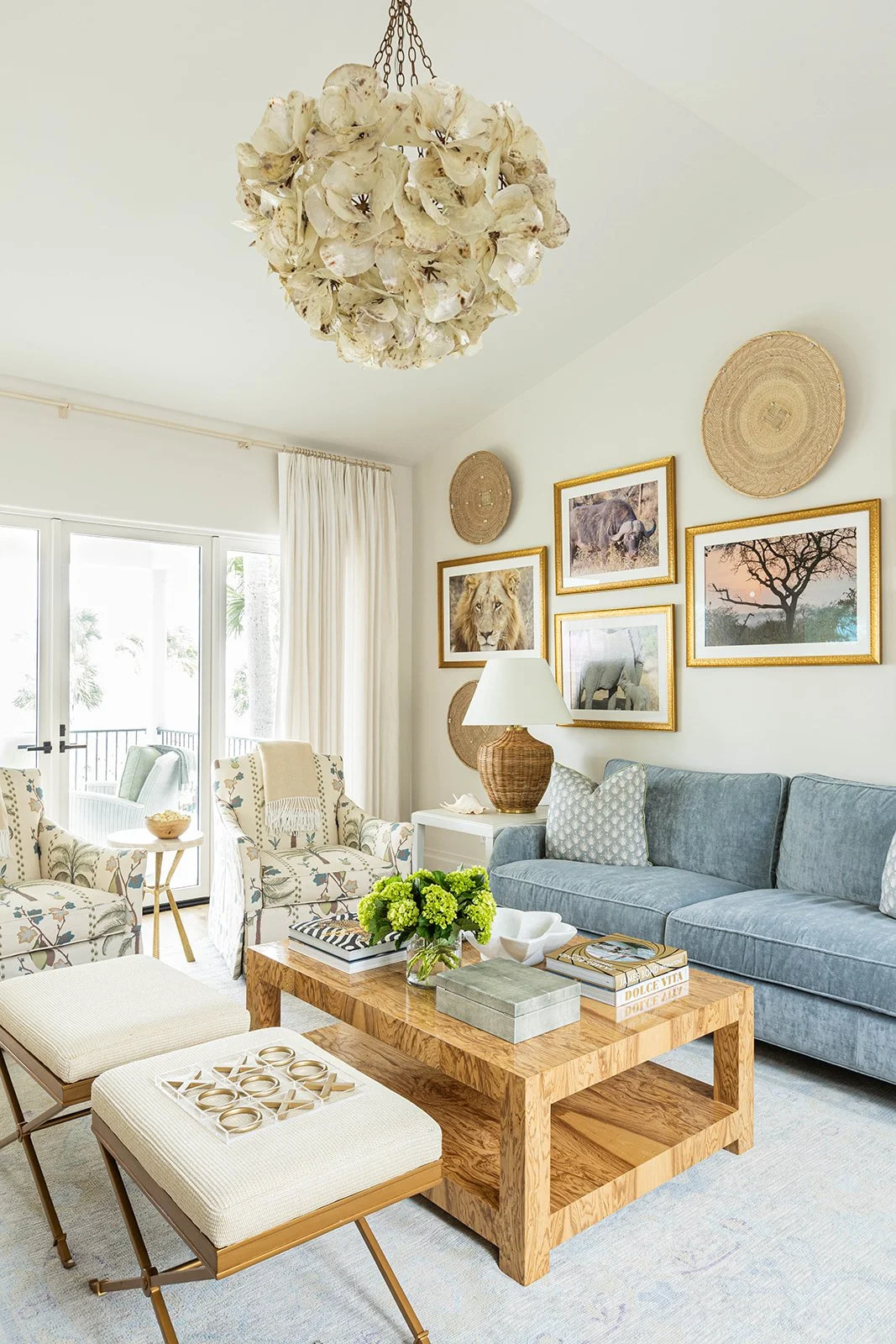 Living room with a chandelier made of shell flowers, framed animal art, wicker wall decorations, a gray sofa with patterned pillows, a wooden coffee table with books and flowers, chairs with patterned fabric, and large glass doors with white curtains.