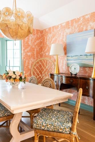 Dining room with coral palm leaf patterned wallpaper, a wooden table with a floral centerpiece, a woven rattan chair, a dark wooden sideboard with a lamp, a clock, and a beach landscape painting, and a large window with green trim.