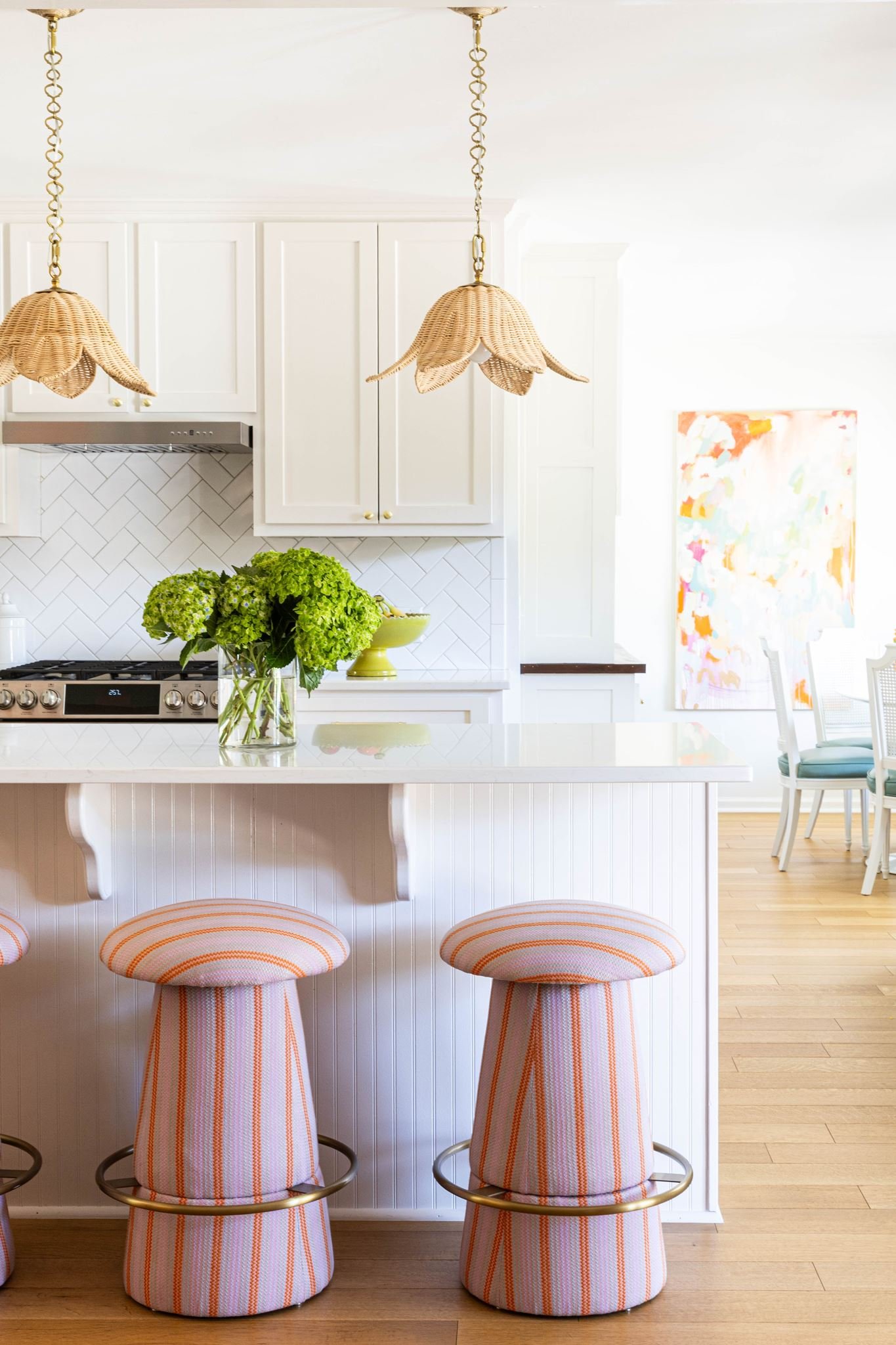 Kitchen with white cabinets, geometric backsplash, stainless steel range hood, two woven pendant lights with floral design, green hydrangea flowers in a vase, yellow cake stand, and a marble island with three striped bar stools.