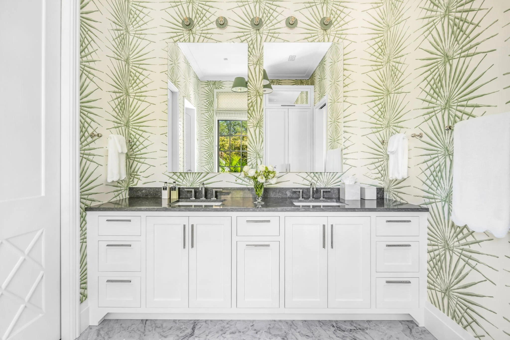 Bathroom with a double vanity, black granite countertop, and white cabinetry, featuring a large mirror, two sinks, and a vase of white roses. The wall behind has tropical leaf-patterned wallpaper, and the room is illuminated by a ceiling light fixture. There are white towels on towel bars and a window showing greenery outside.