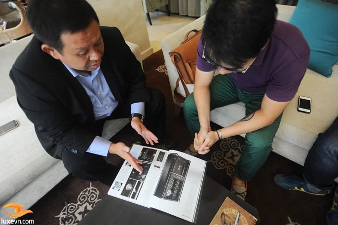 Two men sitting at a small table examining an electronic device catalog or brochure.