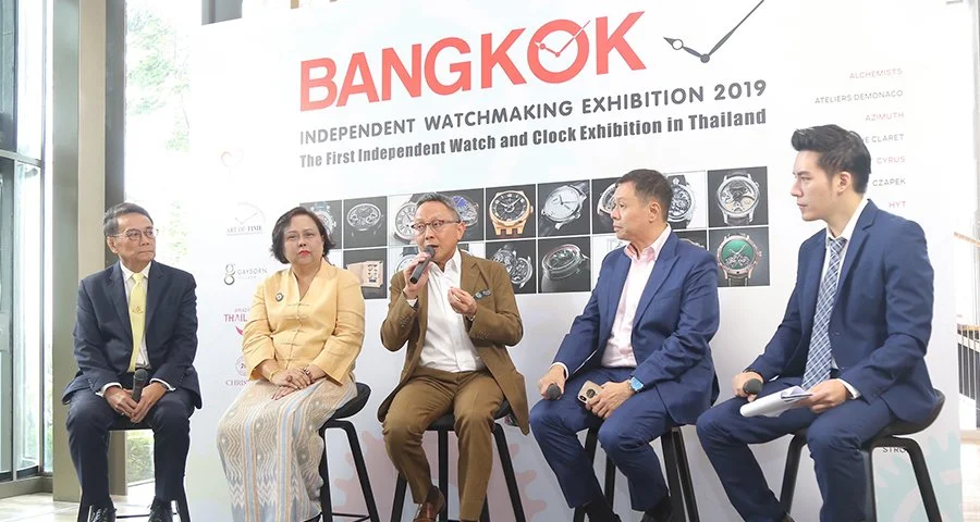 Five people sitting on stools in front of a large board displaying various watches at the Bangkok independent watchmaking exhibition 2019, with a woman and man speaking into microphones.