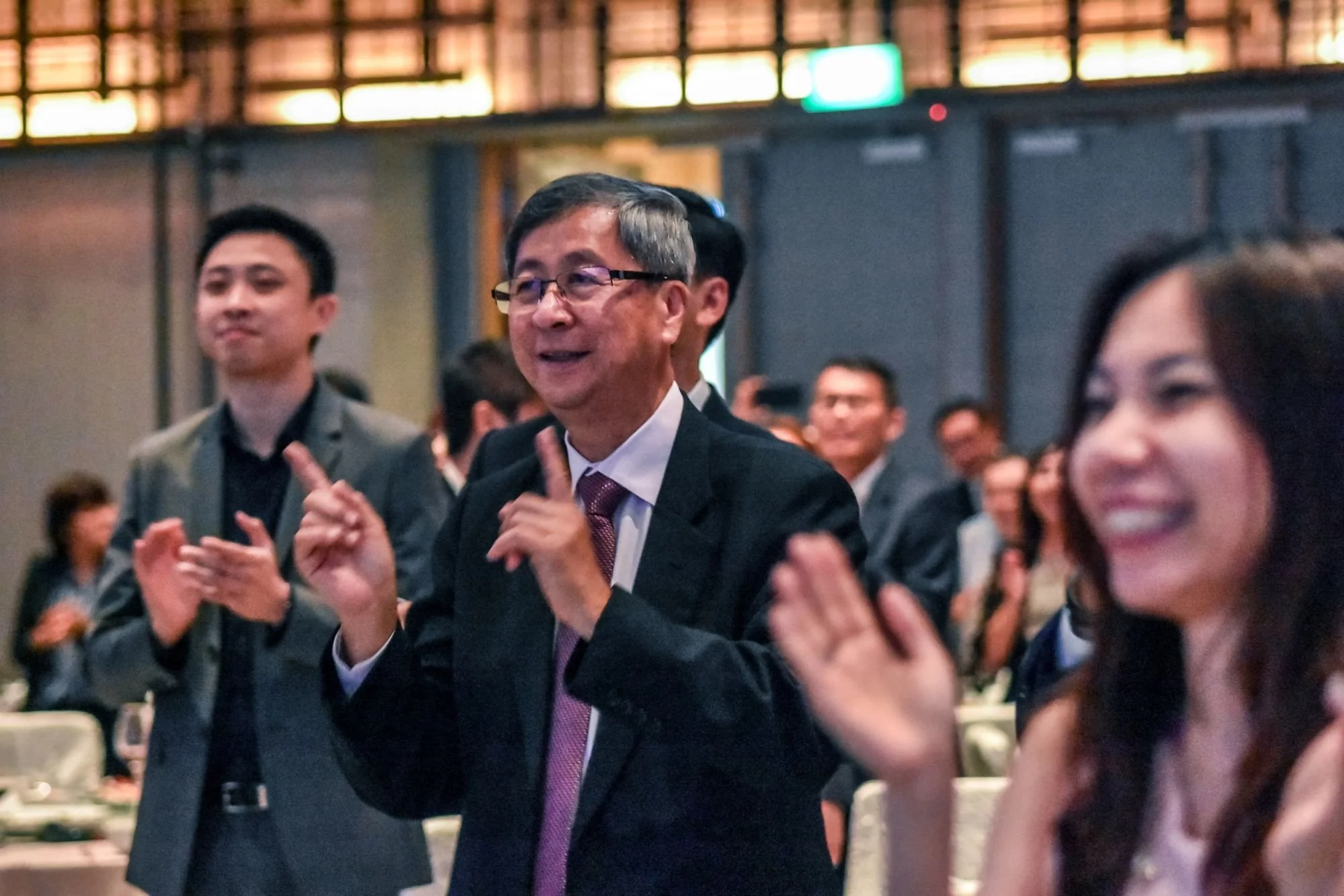People at a formal event clapping and smiling, with a man in the center making gestures with his hands.