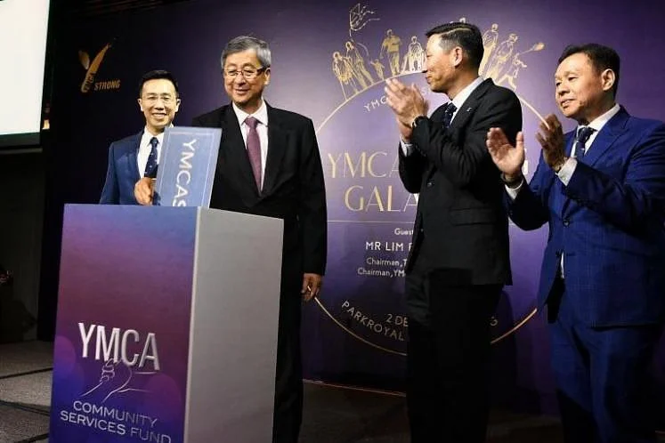 Group of four men in formal attire at a YMCA event, with one man holding a blue folder, standing behind a podium with YMCA Community Services Fund branding, one man clapping, and a purple backdrop featuring event details and illustrations of people.
