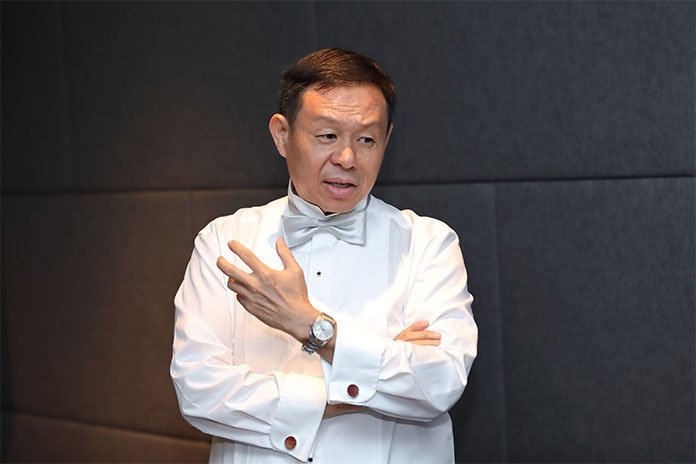 Man dressed as a tuxedo with a bow tie, adjusting his jacket sleeve, standing against a dark wall.