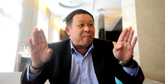 A man in a suit making a stop gesture with both hands, sitting indoors in a well-lit environment.