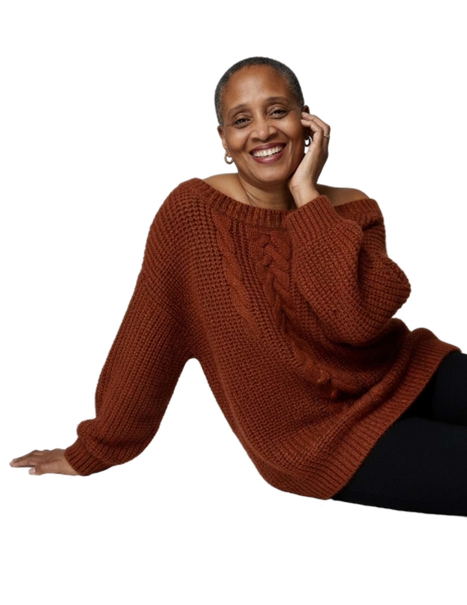 A woman with short hair and brown skin, smiling, wearing a rust-colored knit sweater, sitting with one hand touching her face.