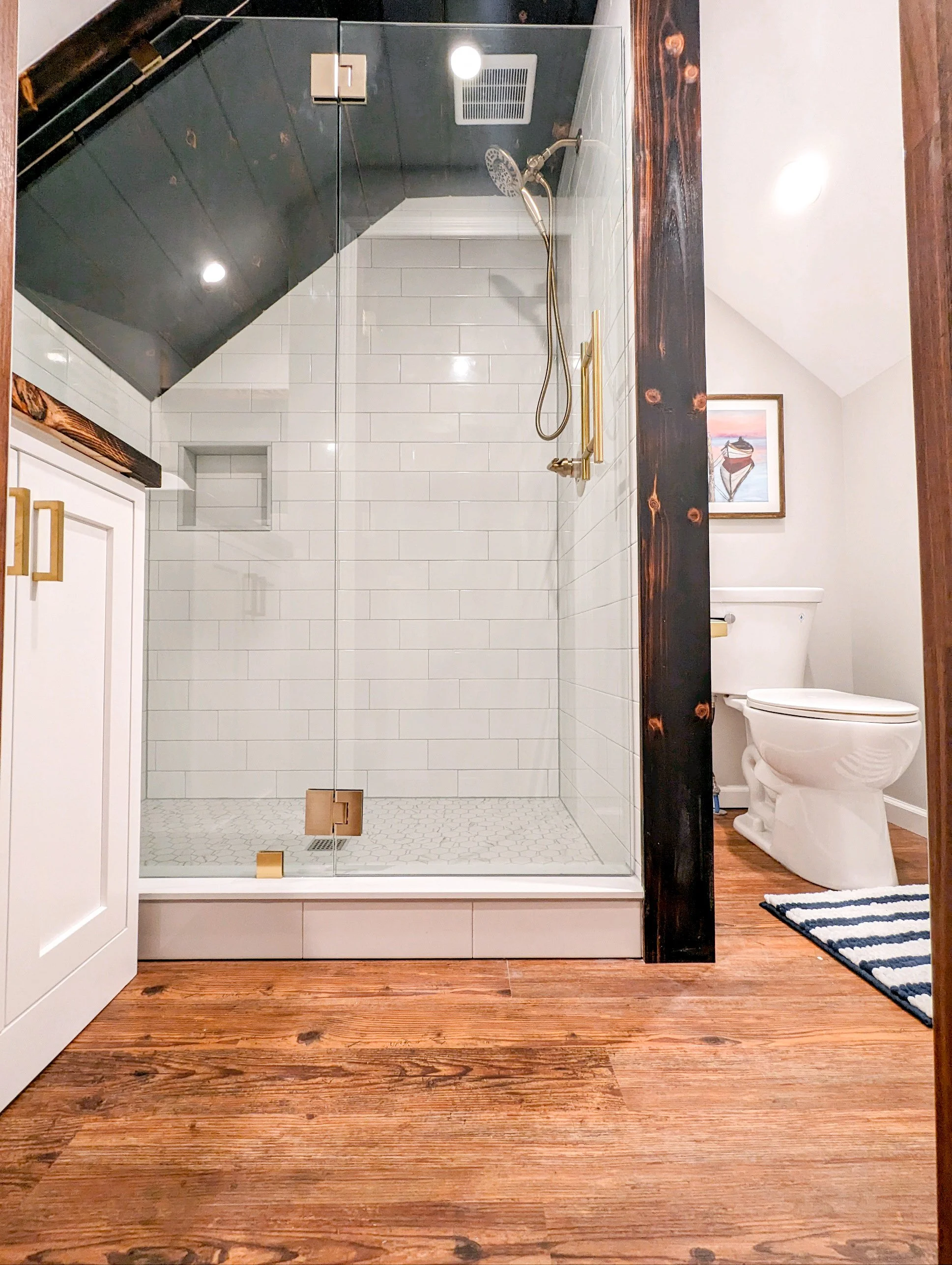 View of a bathroom with a glass-enclosed shower area featuring white subway tiles, a rainfall showerhead, and a niche. To the right, a white toilet is visible, and in the background, a framed painting hangs on the wall. The floor consists of wood planks, and a striped rug is near the toilet.