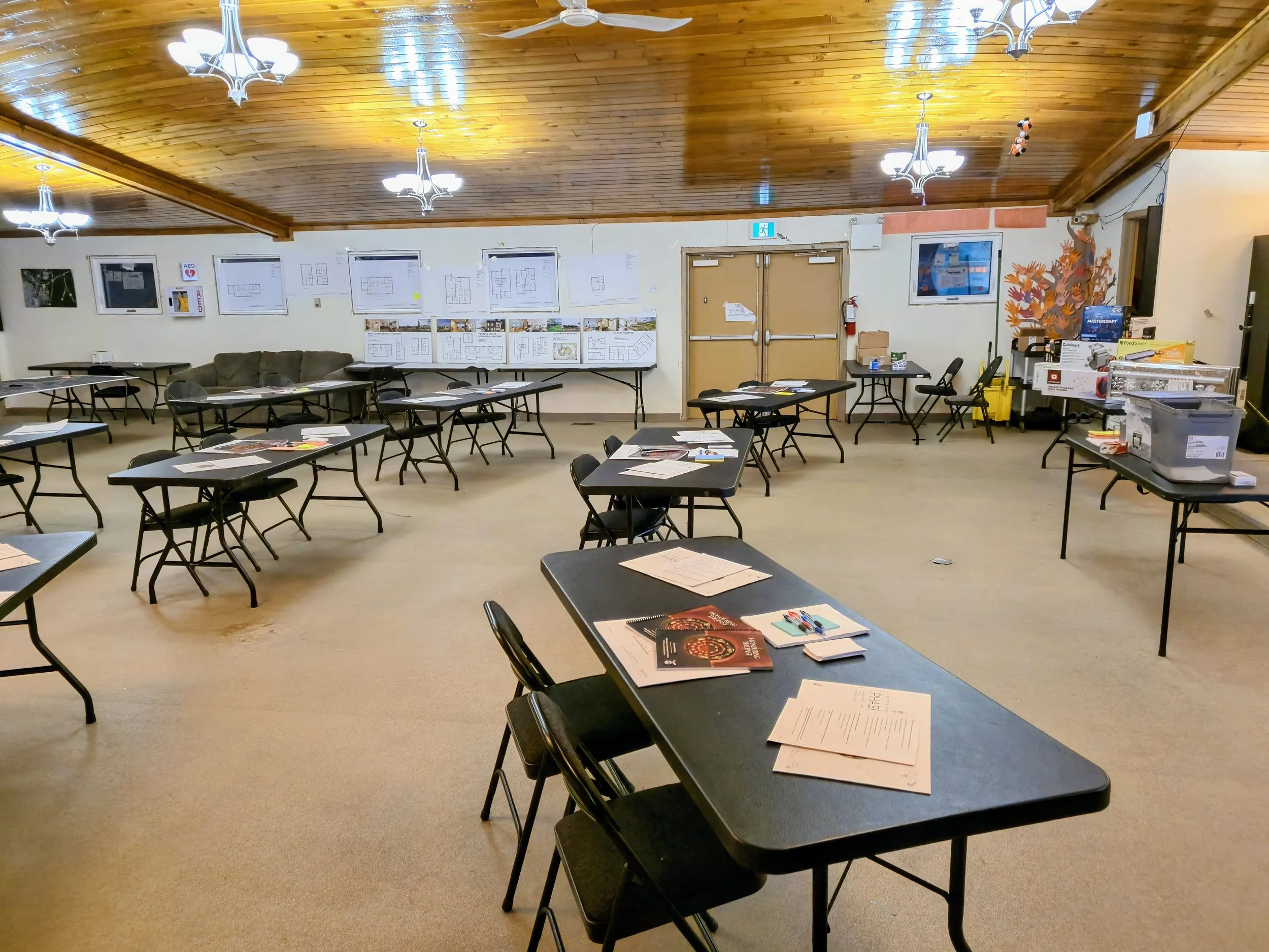 A large room with multiple black tables and chairs, some with papers and pens on them, set up for a meeting or class. The room has a wooden ceiling with hanging light fixtures, white walls decorated with papers and presentation boards, and a few framed posters. There is a door at the back and some storage or refreshment area on the right.