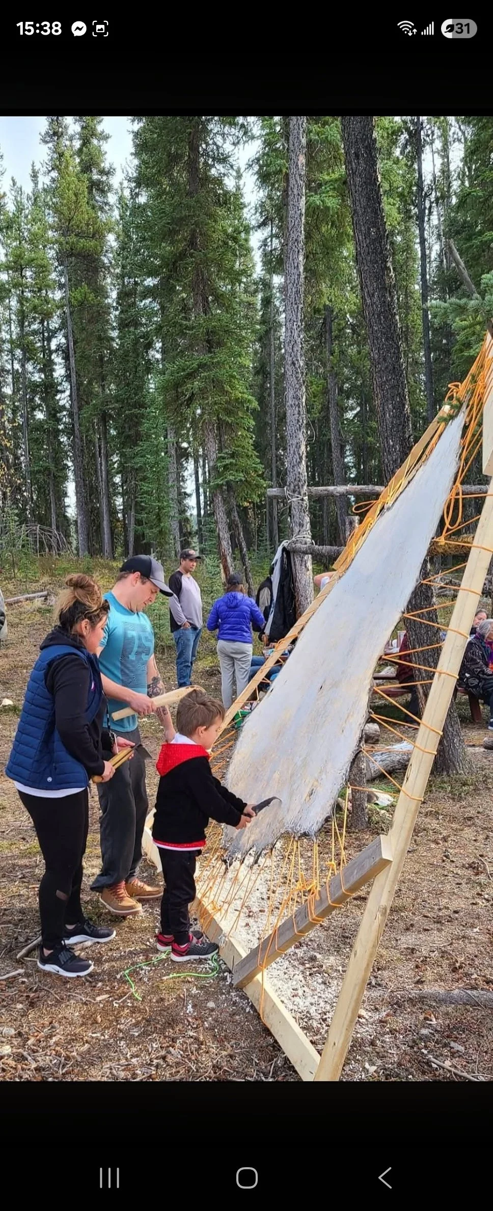 Group of people, including children, engaging in a woodworking activity outdoors in a forested area. They are shaping and working on a large piece of wood secured on a wooden frame.