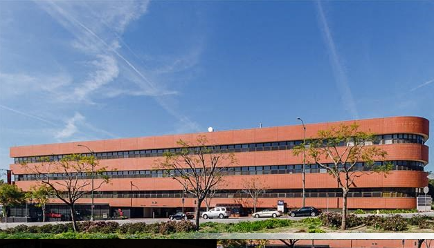 A multi-story office building with orange-brown exterior, surrounded by trees and parked cars, under a blue sky with some clouds.