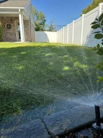 Lawn being watered with a sprinkler in a backyard with a white privacy fence, house, and trees in the background.