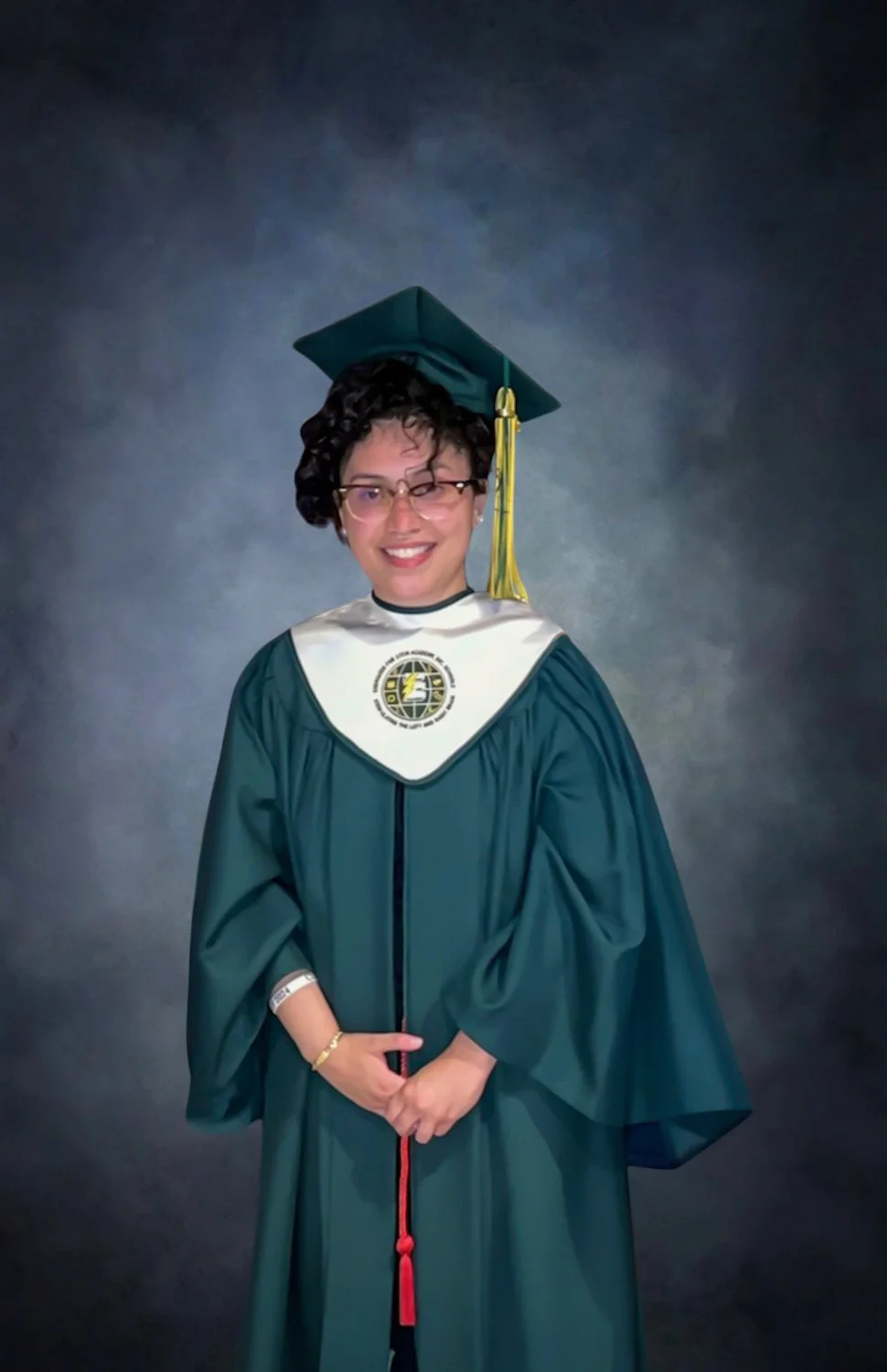 Person wearing a green graduation cap and gown with a white stole and tassel, posing against a dark background.