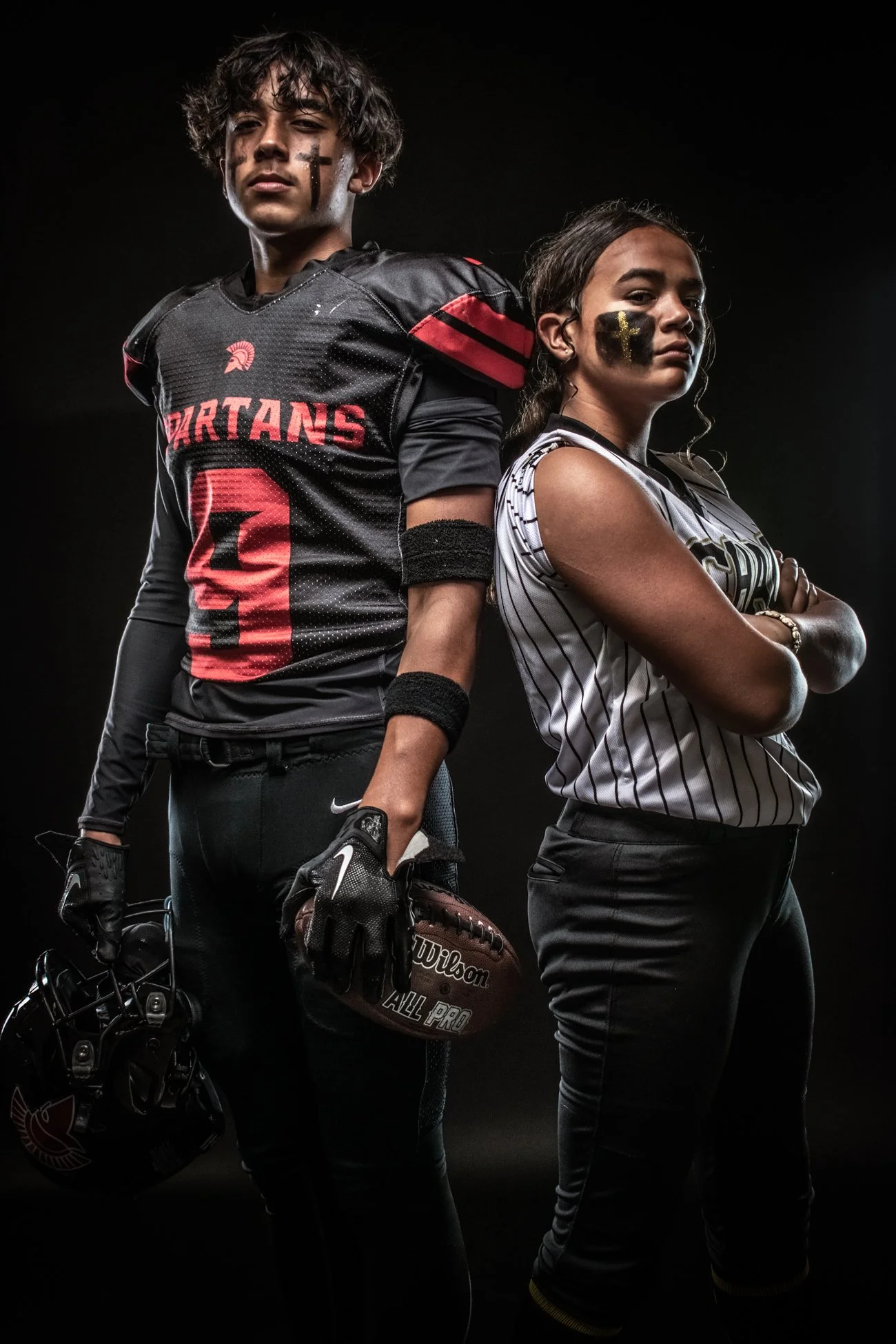 Two young athletes, a male football player in a black and red uniform holding a football, and a female player in a striped sports jersey, standing back to back with arms crossed, against a dark background.