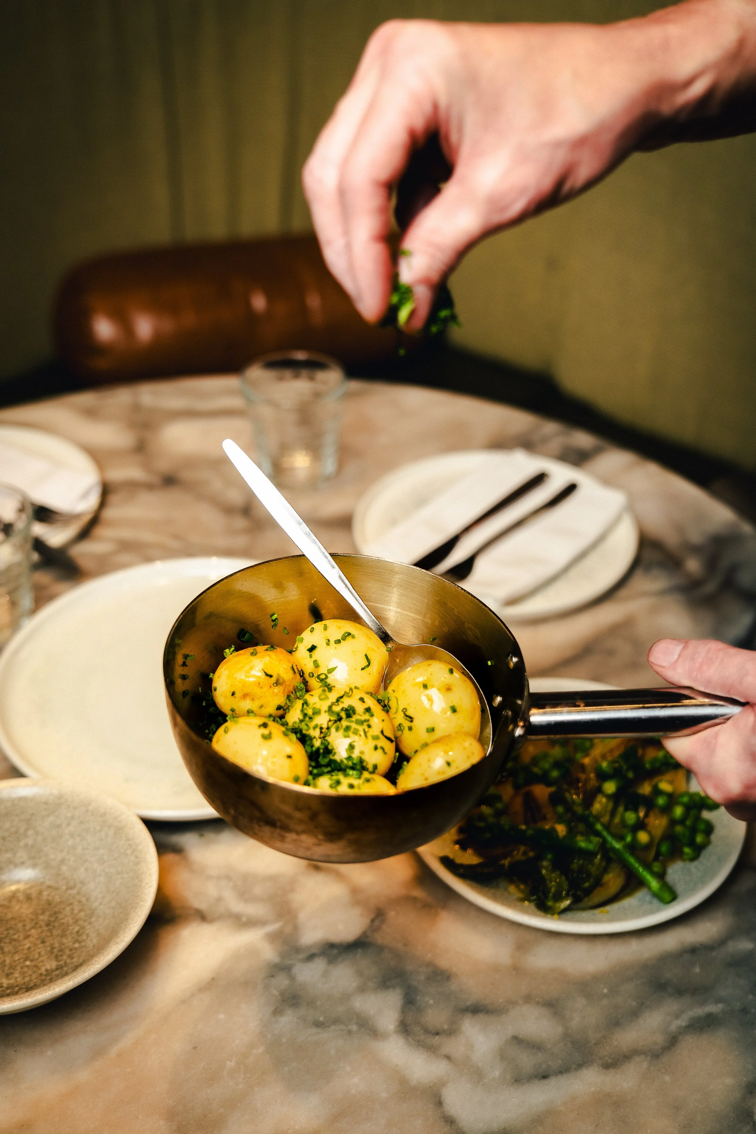 A person holding a small pot of peeled, boiled potatoes garnished with chopped herbs over a marble table with plates, silverware, and bowls.