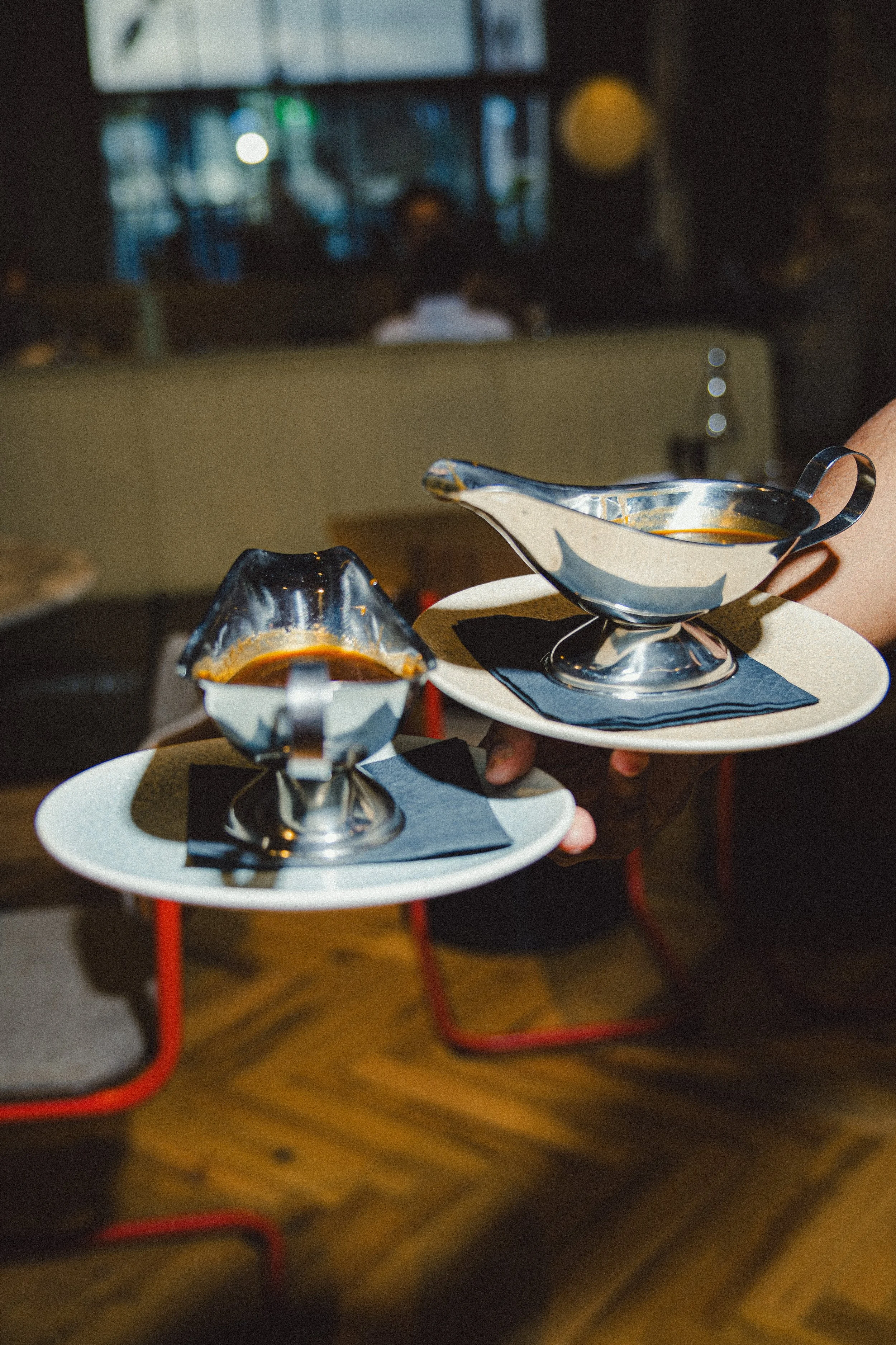 Two people holding trays with silver gravy boats filled with sauce or gravy, served on white plates with black napkins in a restaurant setting.