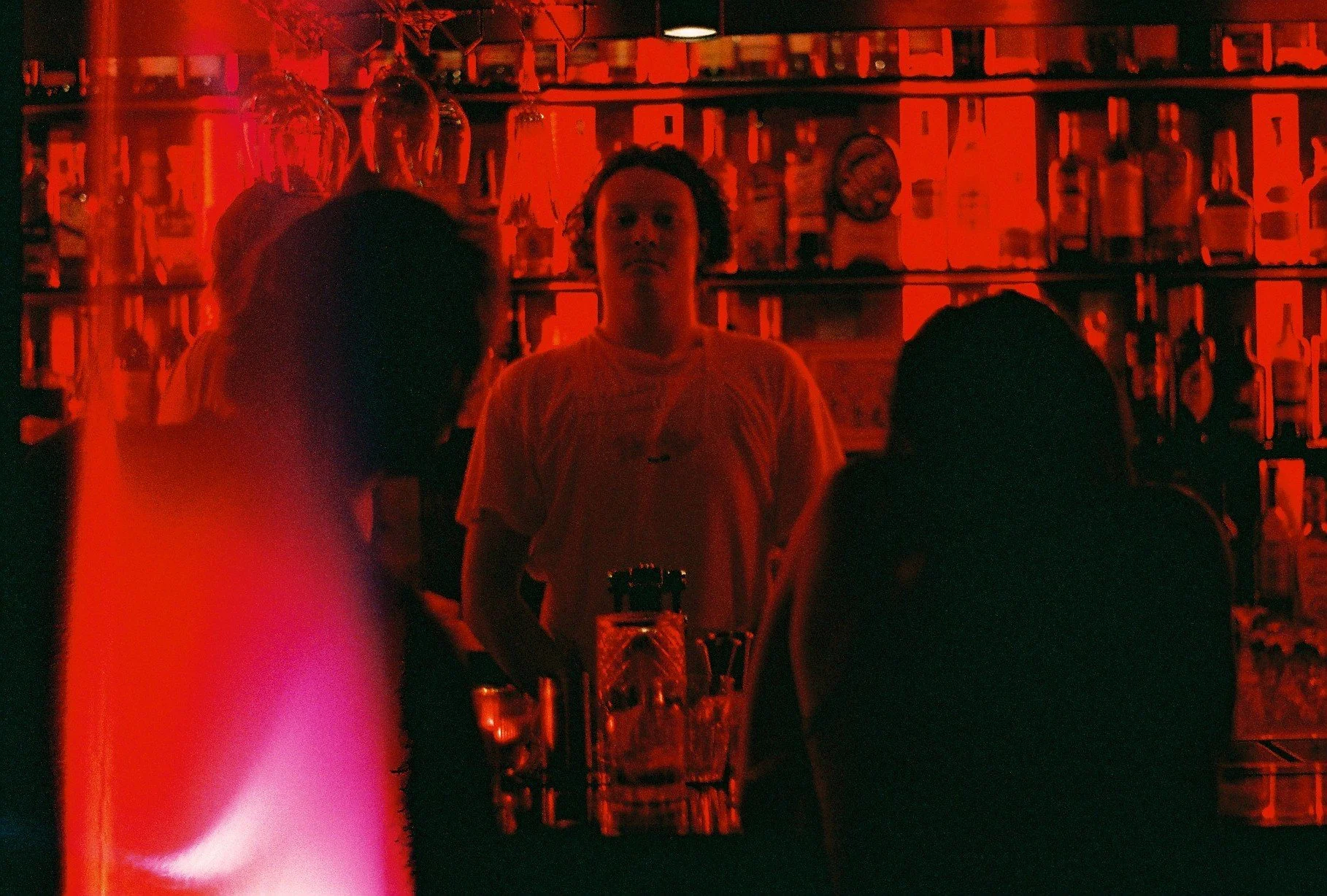 A bartender serving drinks behind a bar with bottles on shelves, lit with red lighting, with three silhouettes of customers in front of the bar.
