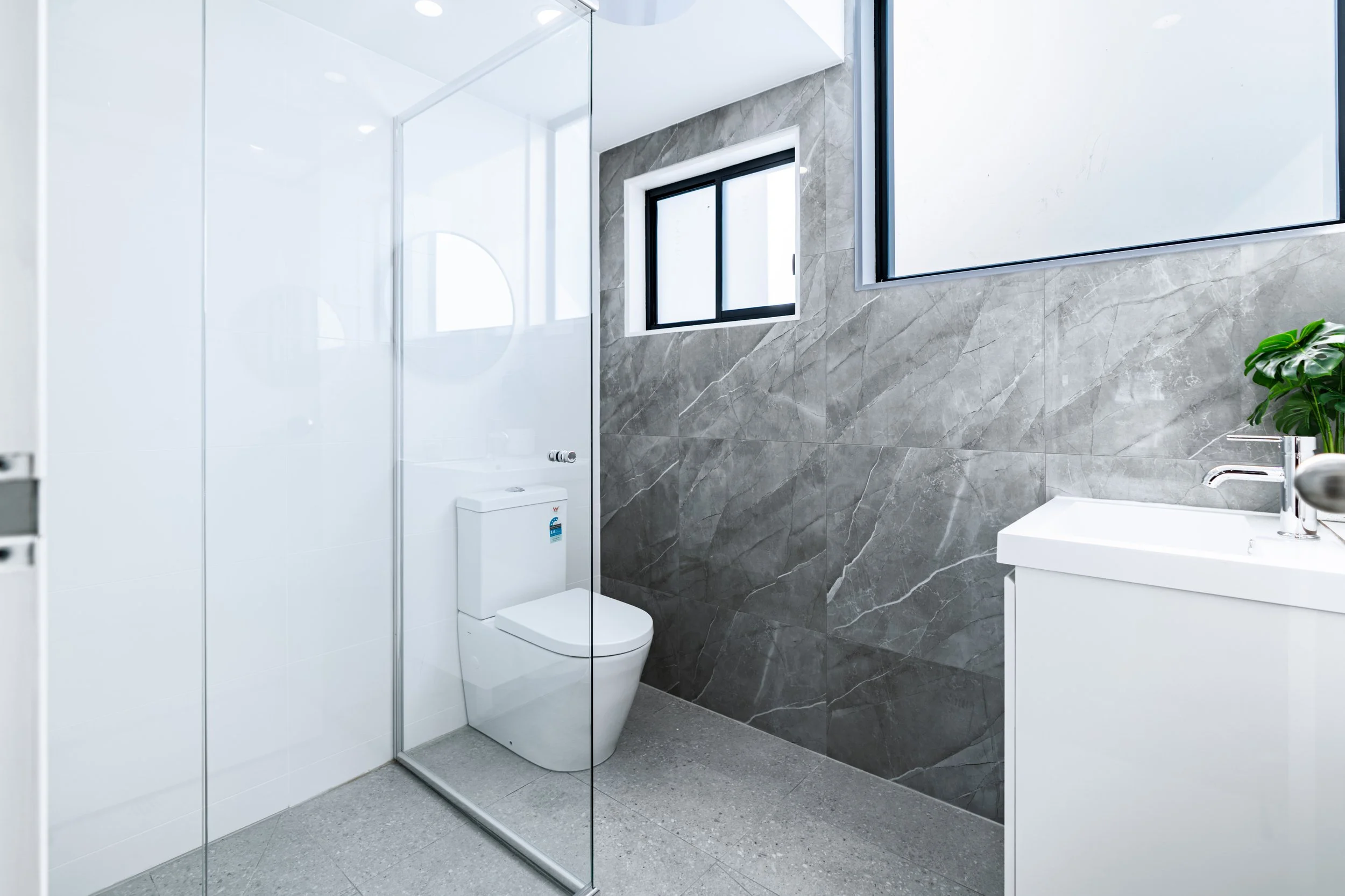 Modern bathroom with gray marble wall, white toilet, glass shower with white panels, small window, white vanity with a green plant, and gray tiled floor.