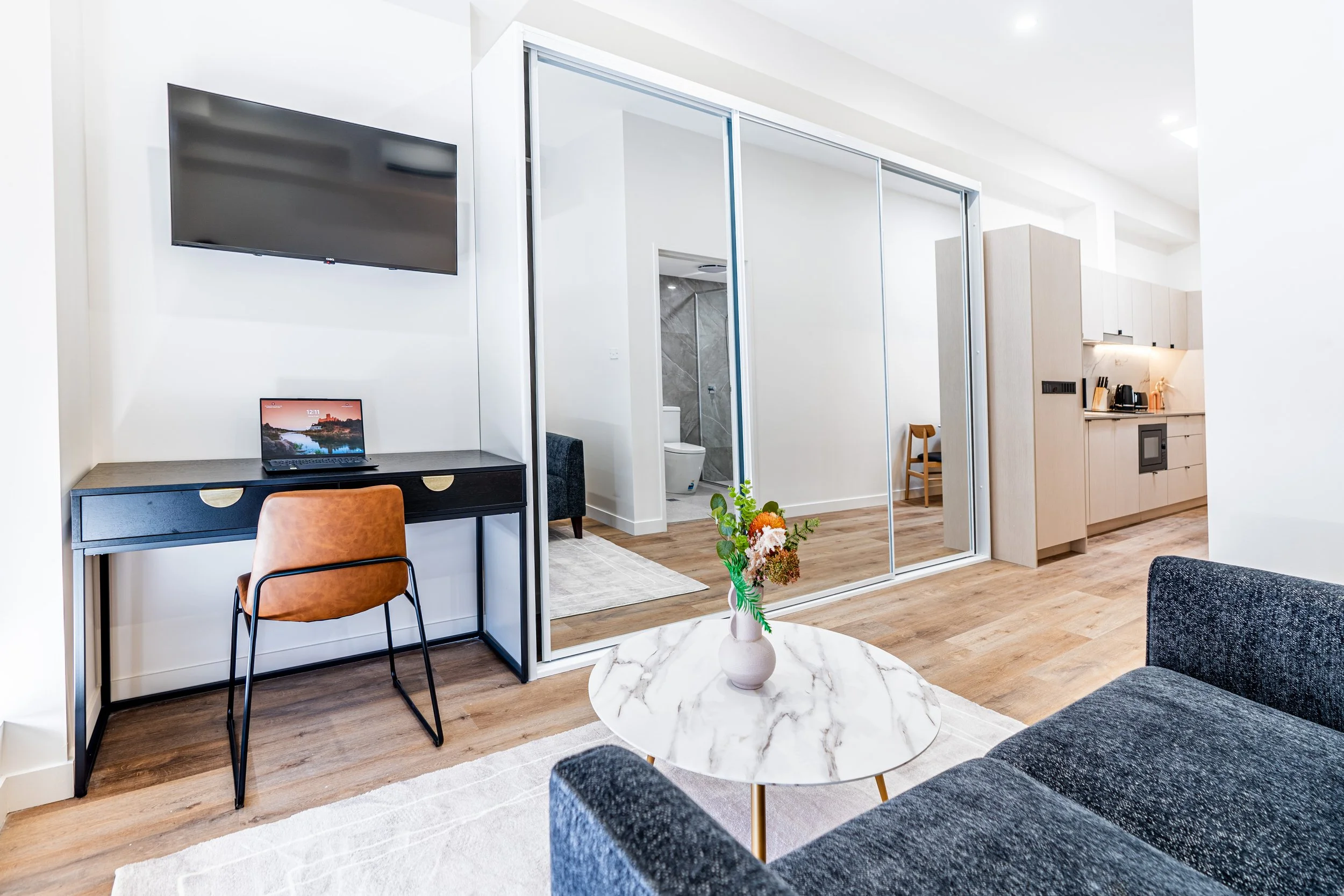 Living room with a black desk and a tan chair, a wall-mounted TV, a marble coffee table with a flower vase, a gray sofa, and a kitchenette with light wood cabinets, a small refrigerator, coffee maker, and oven. A mirrored sliding door reveals a bathr