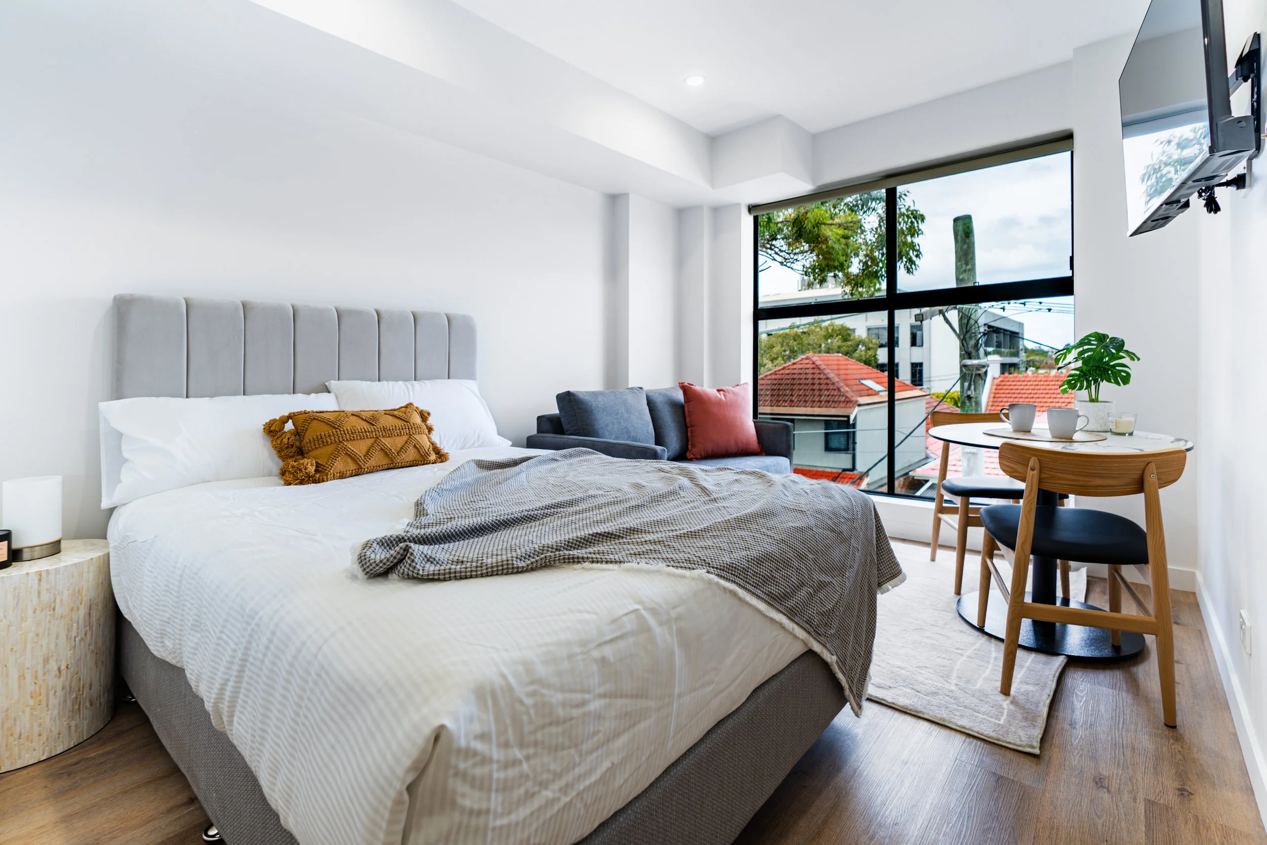 Modern bedroom with a bed, a small round table, a window overlooking rooftops and trees, a TV on the wall, and a small gray sofa with red and brown cushions.