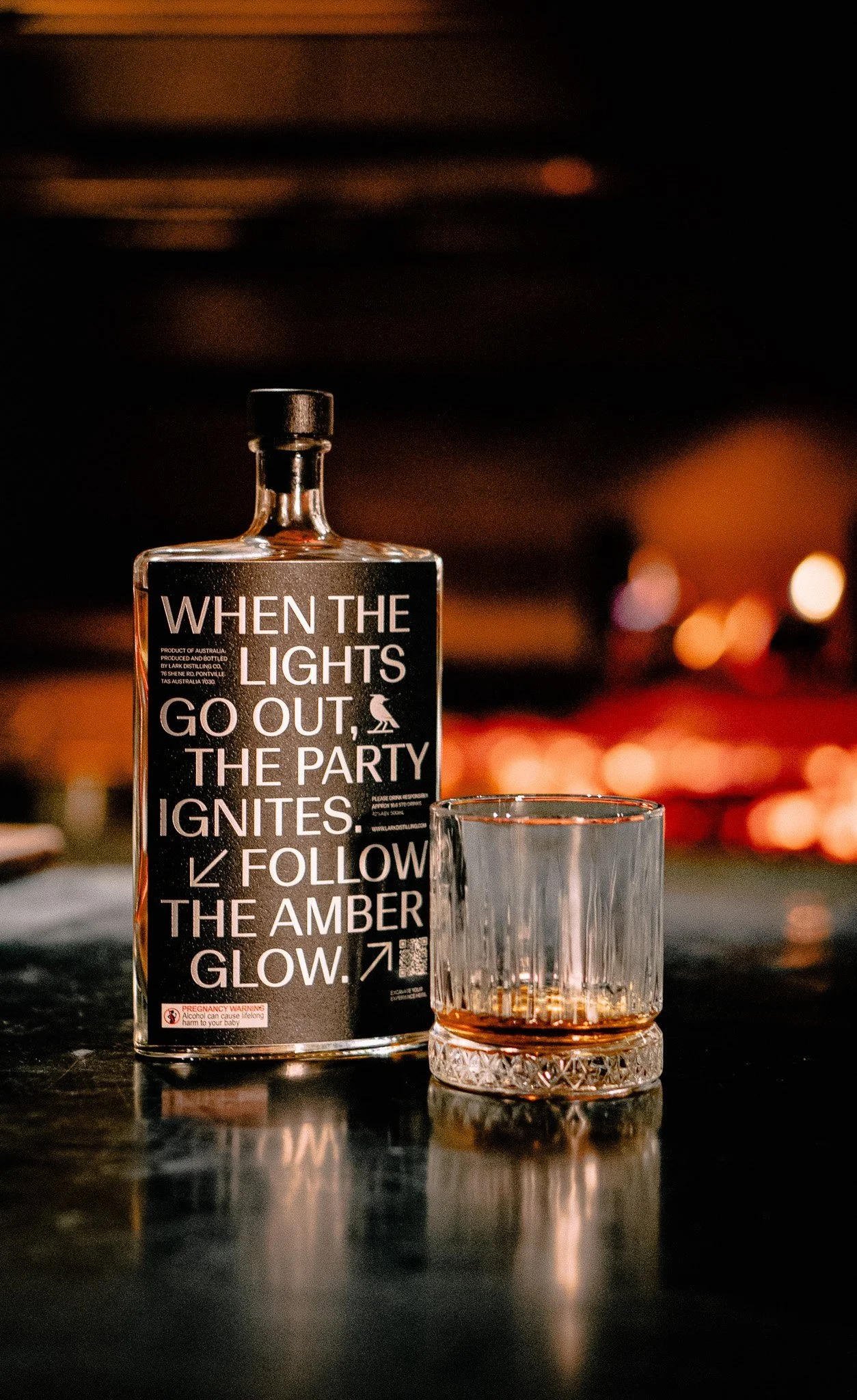A bottle with a black label and white text reads, 'When the lights go out, the party ignites. Follow the amber glow.' Next to it is a glass with a small amount of amber-colored liquid inside, on a reflective dark surface with blurred warm lights in the background.